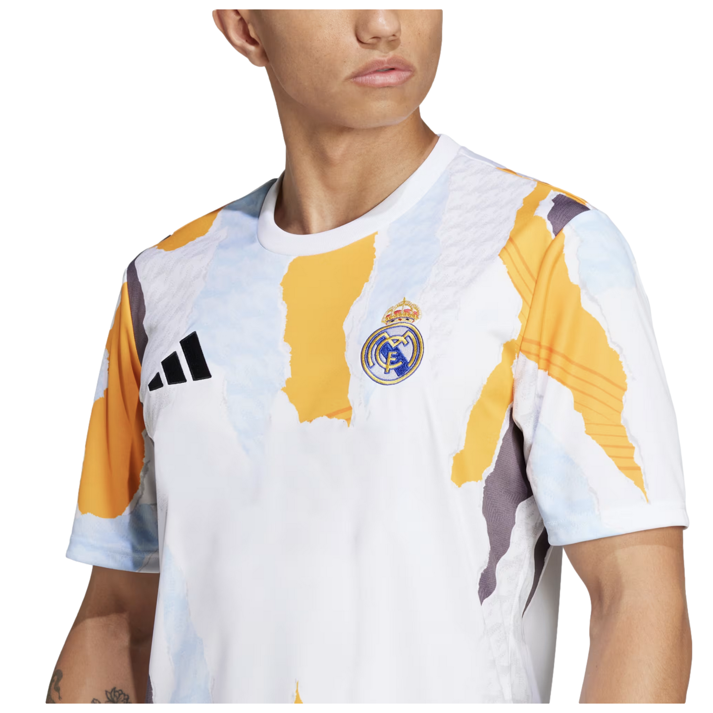 A person wearing an Adidas Real Madrid pre-match jersey featuring a white base with an abstract yellow, light blue, and grey torn-paper pattern. The Real Madrid club crest is visible on the left chest and the black Adidas logo on the right.