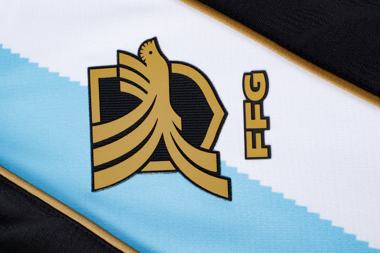 A stylized gold bird logo with vertical "FFG" text on a white and light blue patterned sports jersey with black and gold trim.