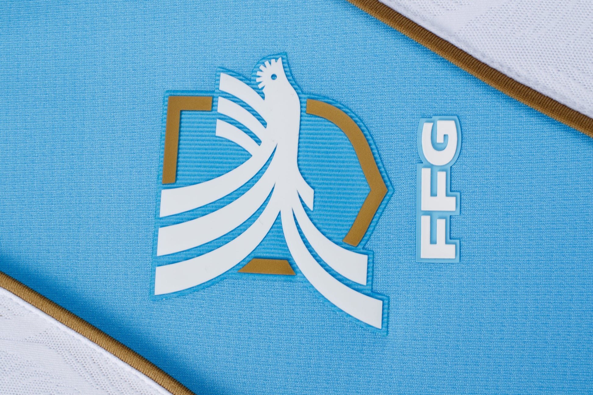 Close-up of a white rooster logo with gold accents and "FFG" text on light blue athletic fabric. White and gold trim is visible at the corners.