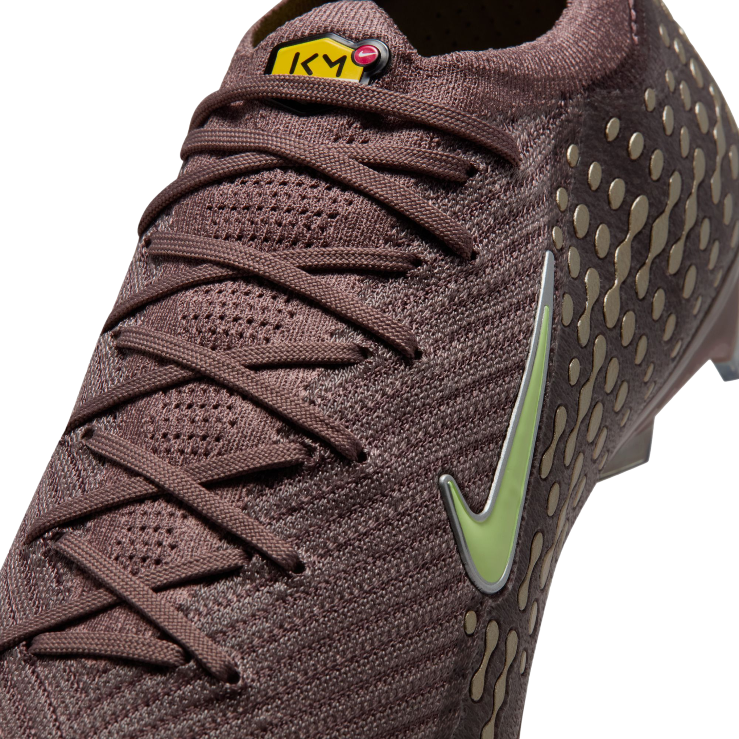 Close-up of a mauve Nike soccer cleat with a knit texture, lime green swoosh, and yellow KM logo on the tongue. Gold dot patterns are visible on the side.