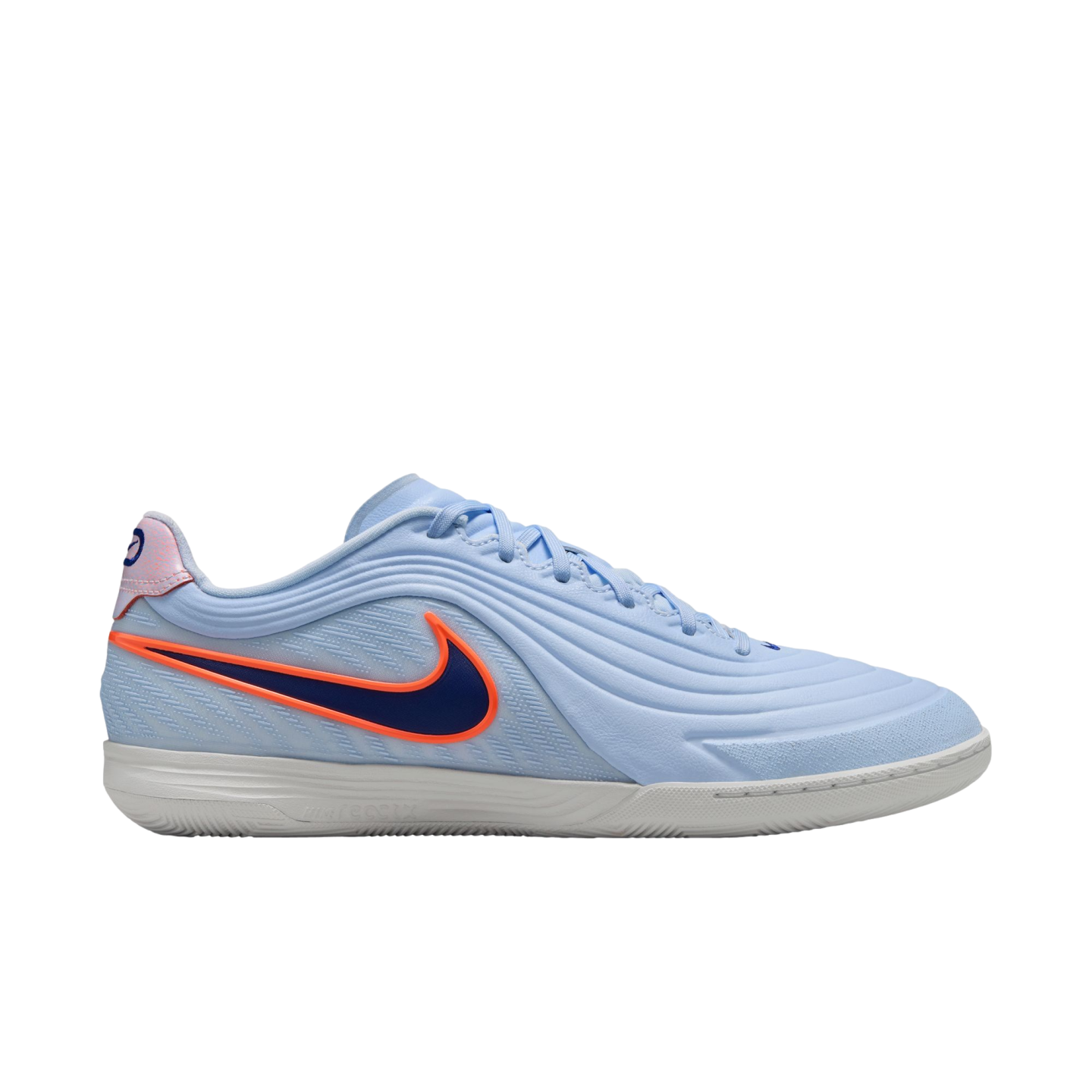 A side-view profile of a light blue Nike indoor soccer shoe against a black background. The shoe features a textured upper with wavy lines, a dark blue Swoosh with a bright orange outline, and a thin, white rubber sole.