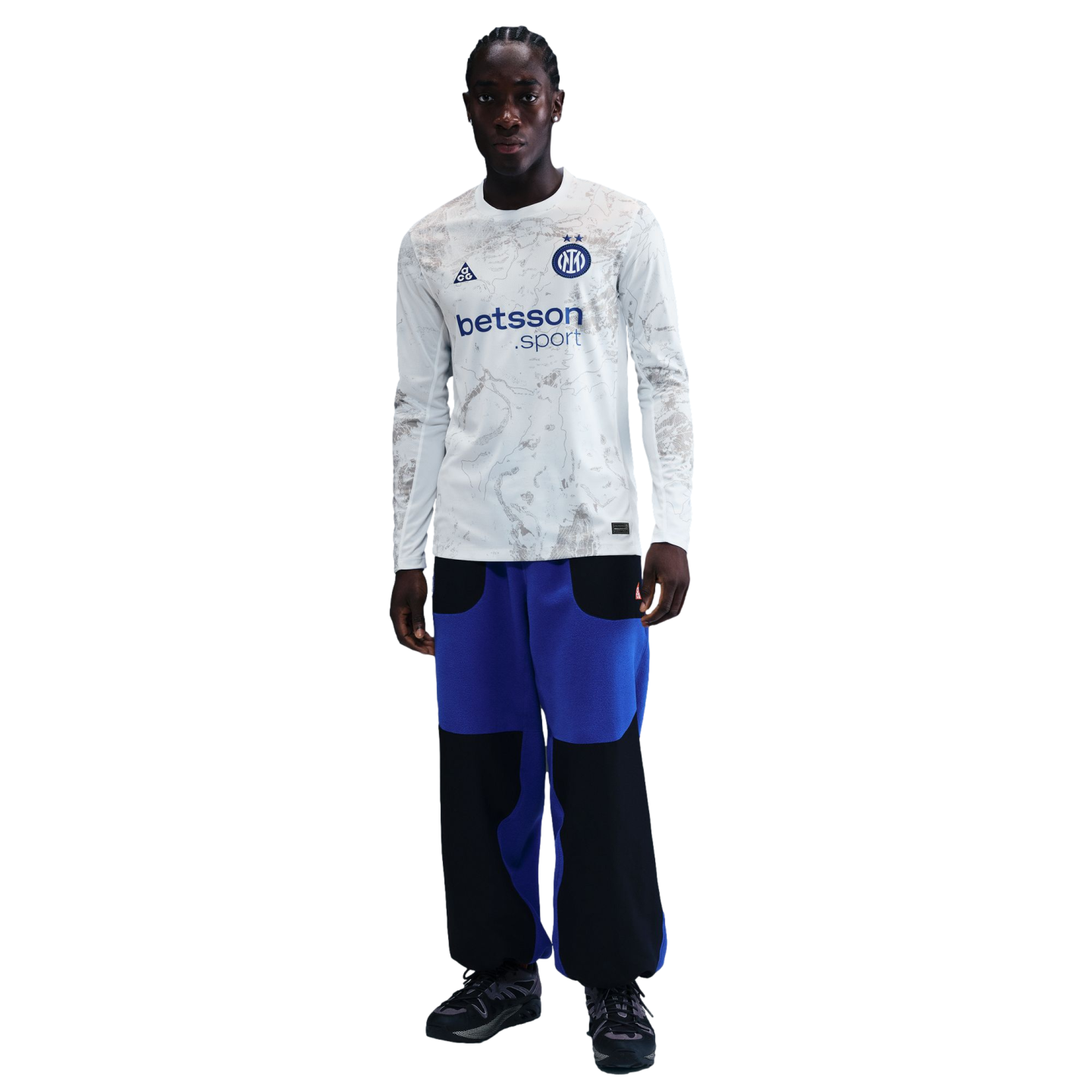 A young Black man with braids wears a long-sleeve white jersey with gray patterns and a "betsson .sport" logo, blue and black pants, and dark sneakers, standing against a black background.
