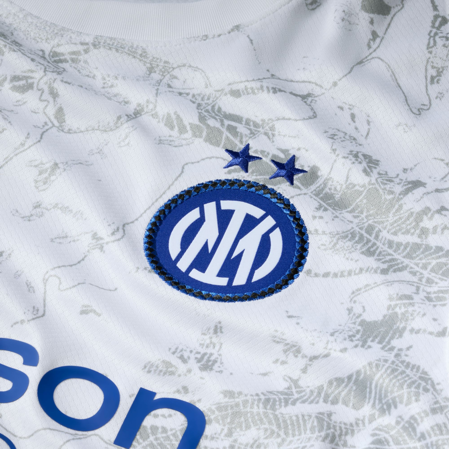 Close-up of a white Inter Milan jersey featuring the blue and white club crest with two stars above it on a subtle grey-patterned fabric.