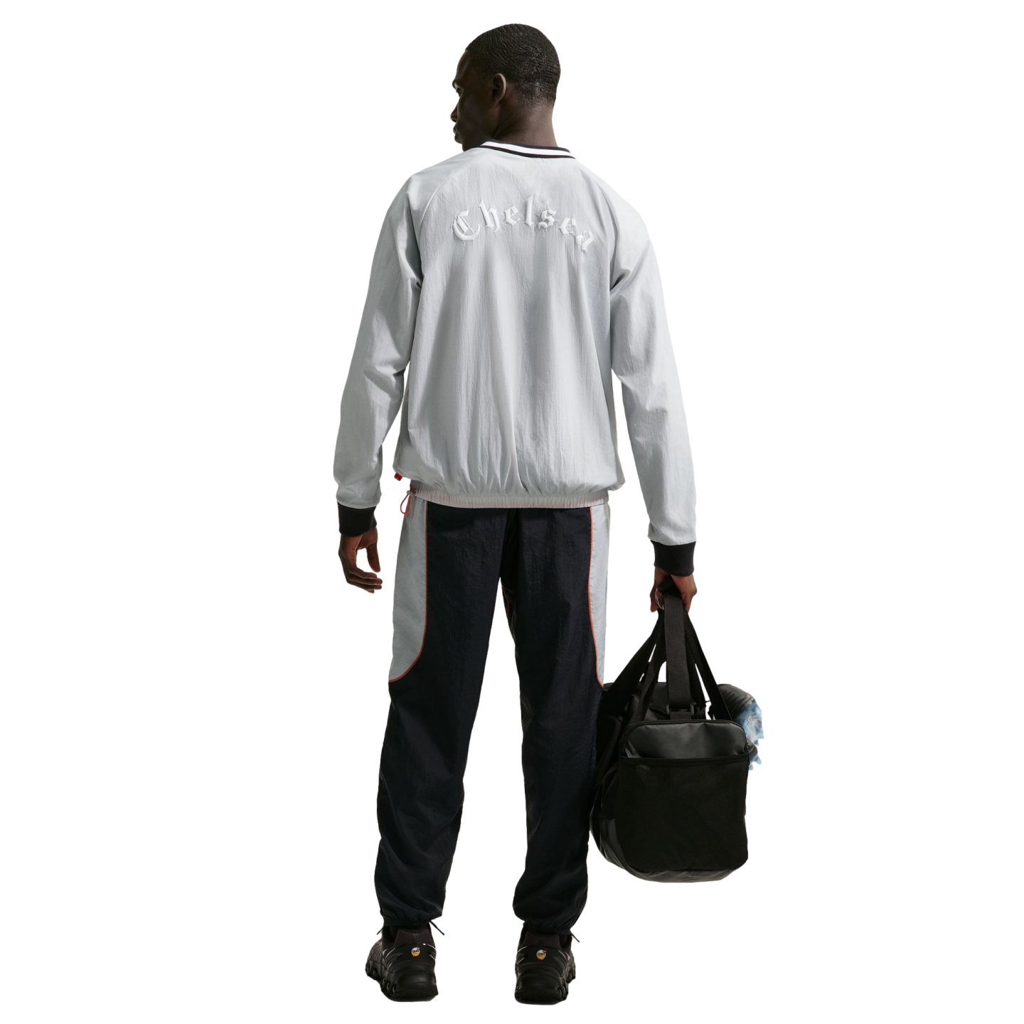 Rear view of a man in a grey 'Chelsea' jacket and navy track pants, holding a black duffel bag against a black background.