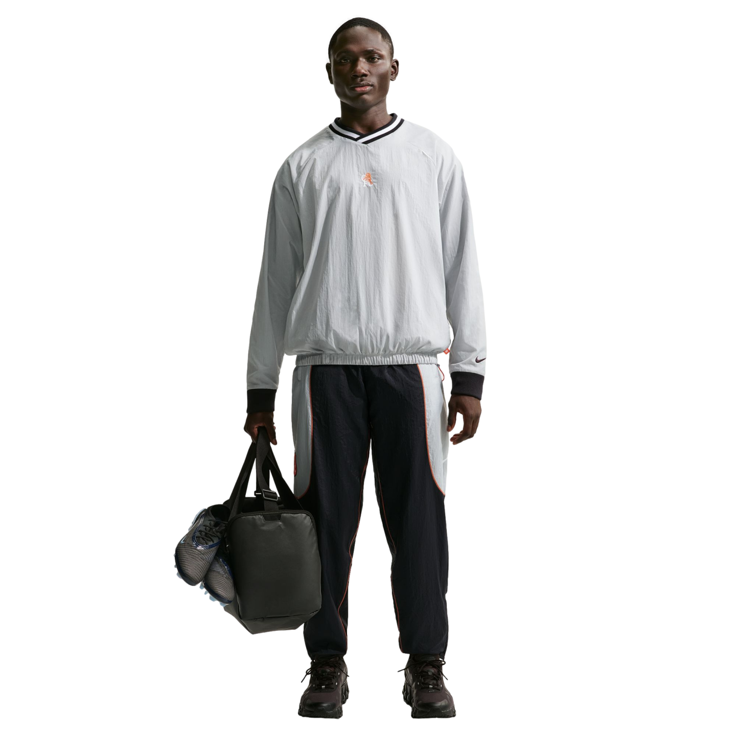 Full-length shot of a man in a grey pullover and navy track pants, holding a black duffel bag with blue cleats against a black background.