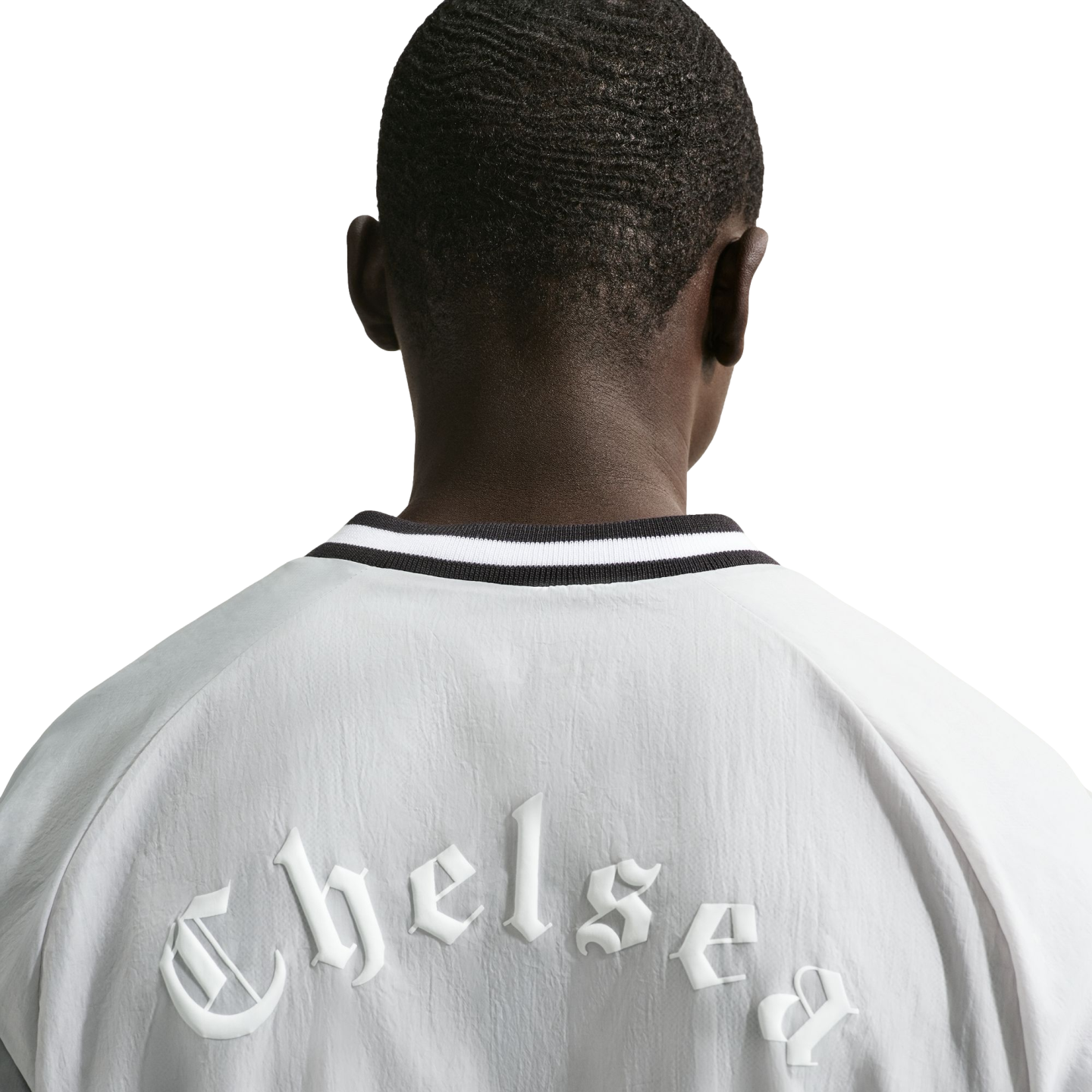 View from behind of a person wearing a white jacket. Across the upper back, the word "Chelsea" is embossed in a white, gothic-style font. The jacket has a black and white striped collar.