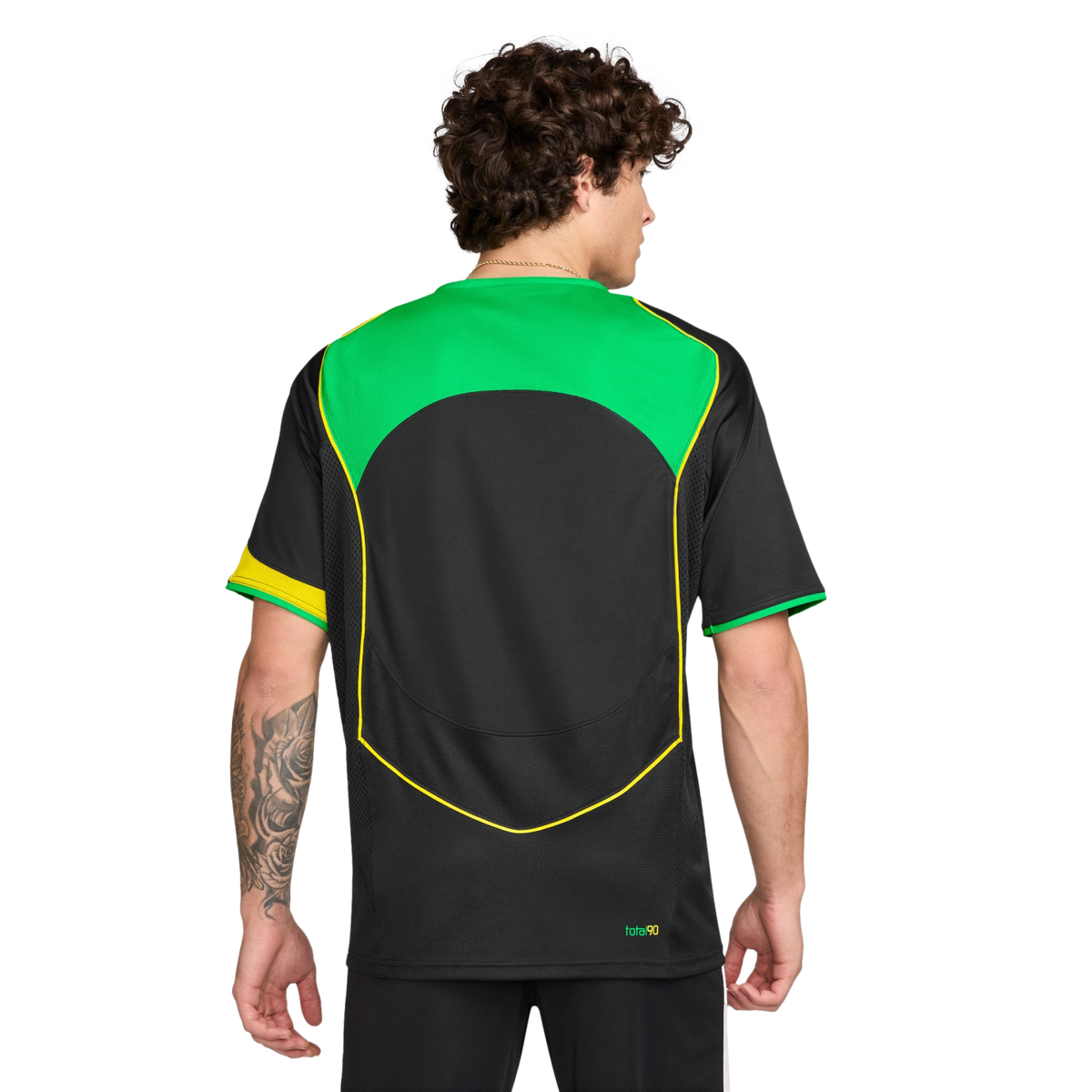 Rear view of a man with curly hair wearing a black soccer jersey with green and yellow accents. He has a tattoo on his left forearm. Plain white background.
