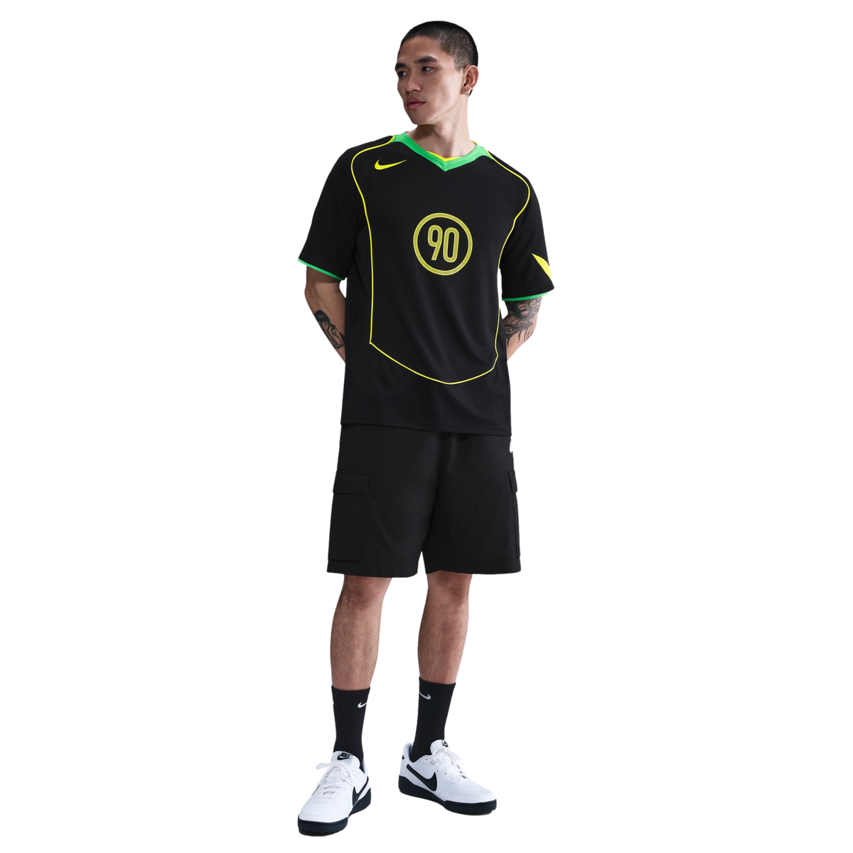 A man poses in a black Nike T90 soccer jersey, black cargo shorts, and white sneakers against a white background.