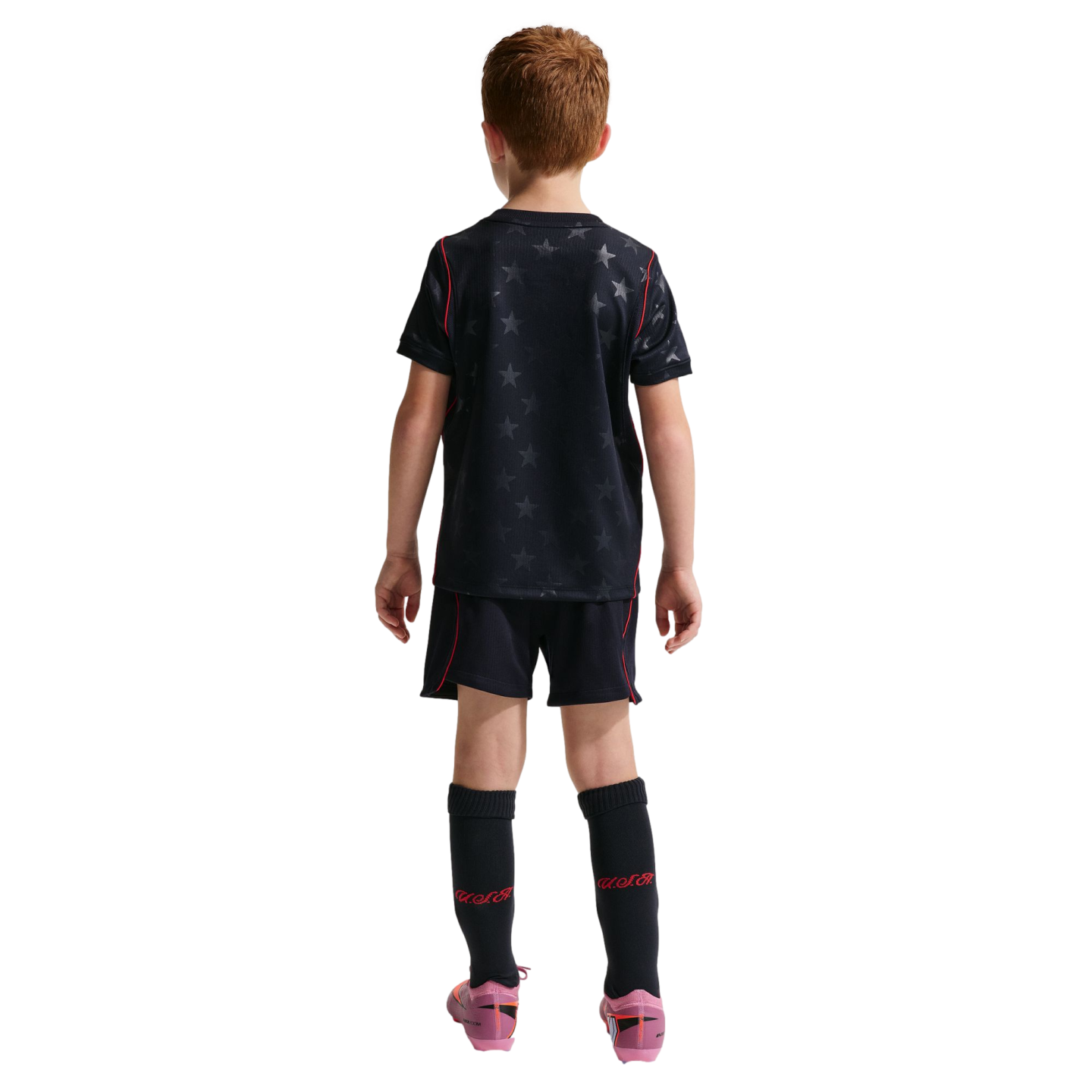 Rear view of a young boy with red hair wearing a black soccer kit with a star pattern and red trim. He is standing against a solid black background, wearing tall black socks with "U.S.A." in red and pink soccer cleats.