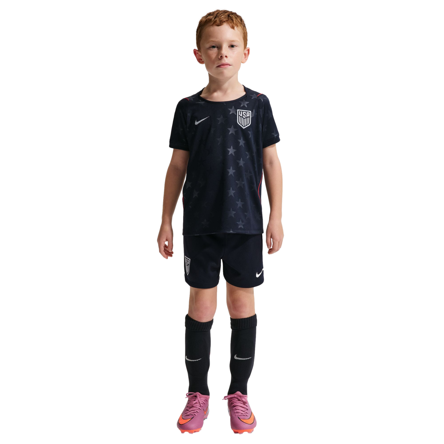 A young boy with red hair standing against a black background, wearing a dark blue Nike USA soccer kit with matching shorts, black socks, and pink cleats.