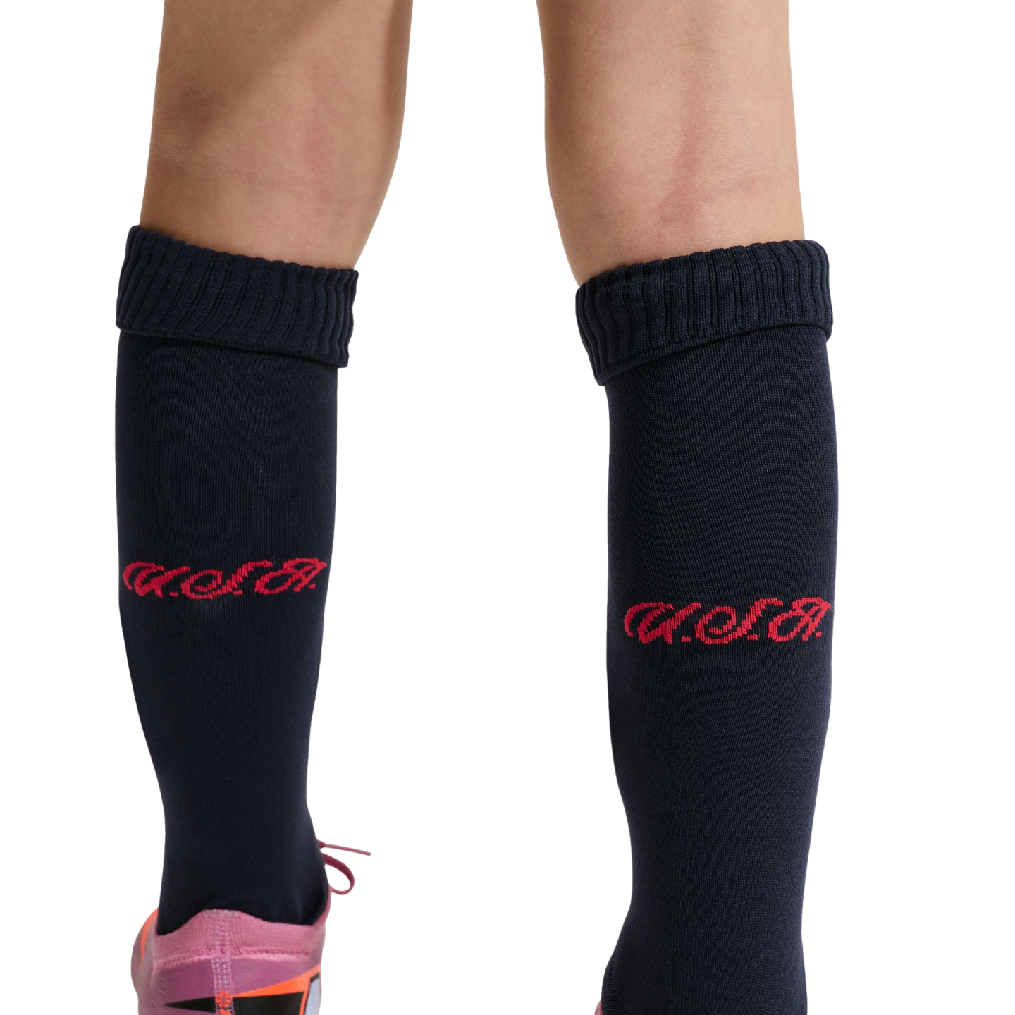 A close-up of a person's lower legs against a black background, wearing navy blue knee-high socks with a red script logo and a glimpse of a pink and orange athletic shoe.