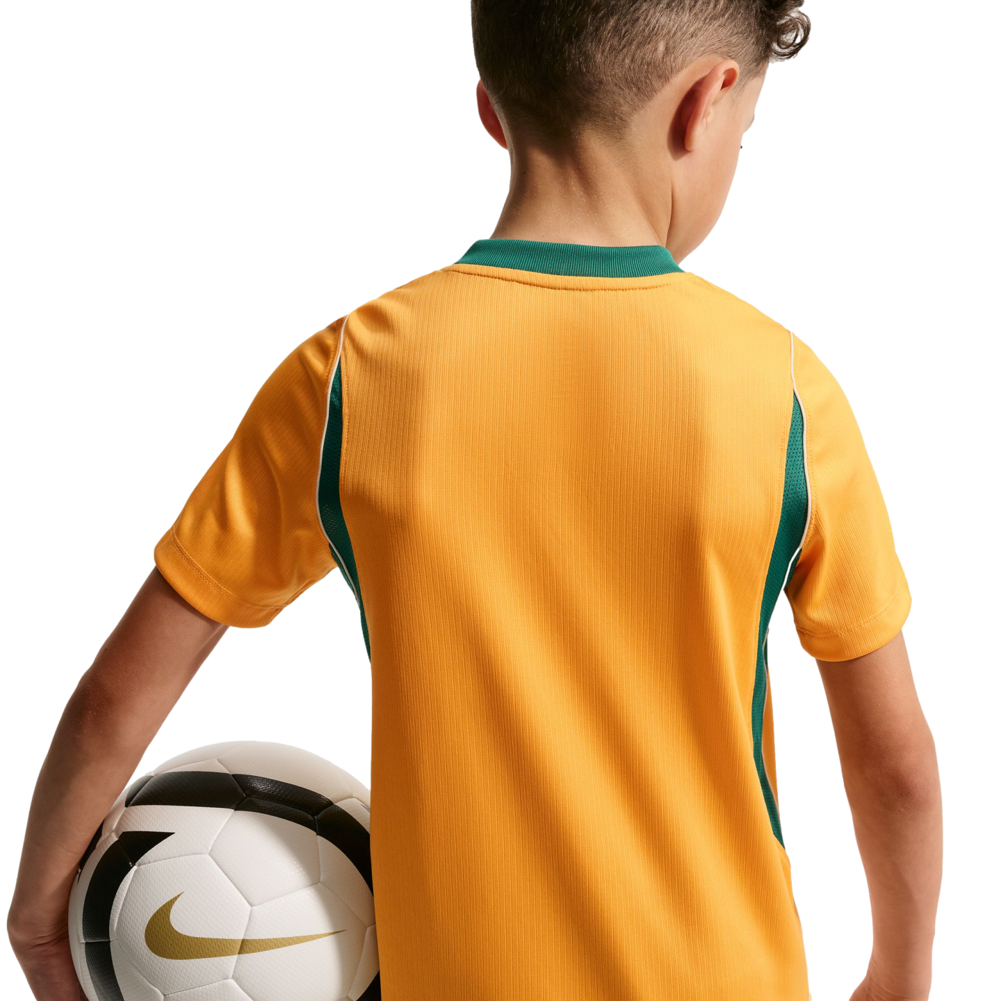 A rear view of a child wearing a gold soccer jersey with green trim, holding a white soccer ball under their arm against a solid black background.