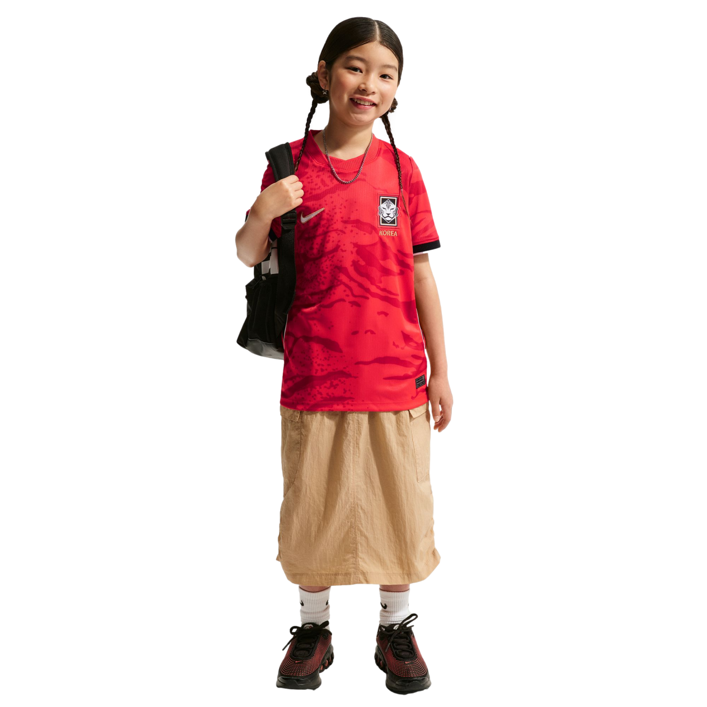A smiling girl with braids wears a red Korea soccer jersey, tan skirt, and black backpack against a black background.
