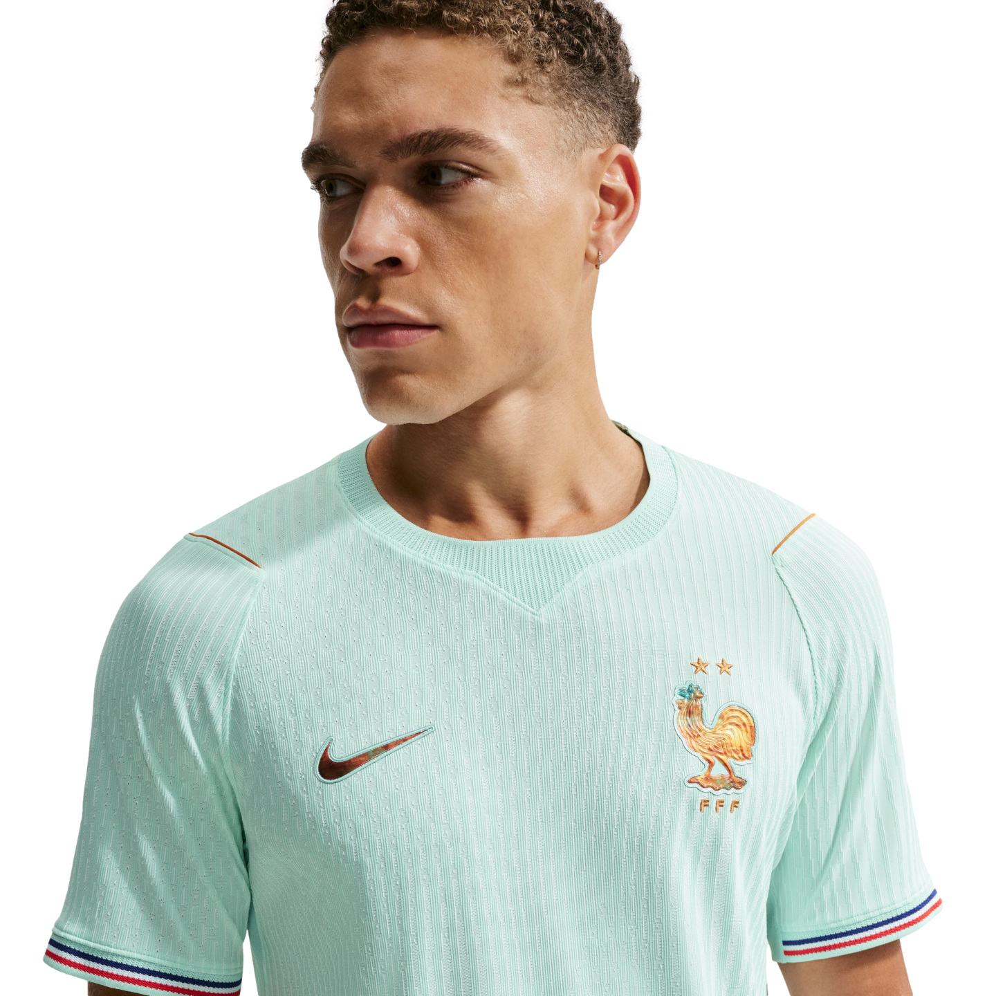 A man wears a mint green France national soccer team jersey with a golden rooster crest and tricolor sleeve cuffs, looking away against a black background.