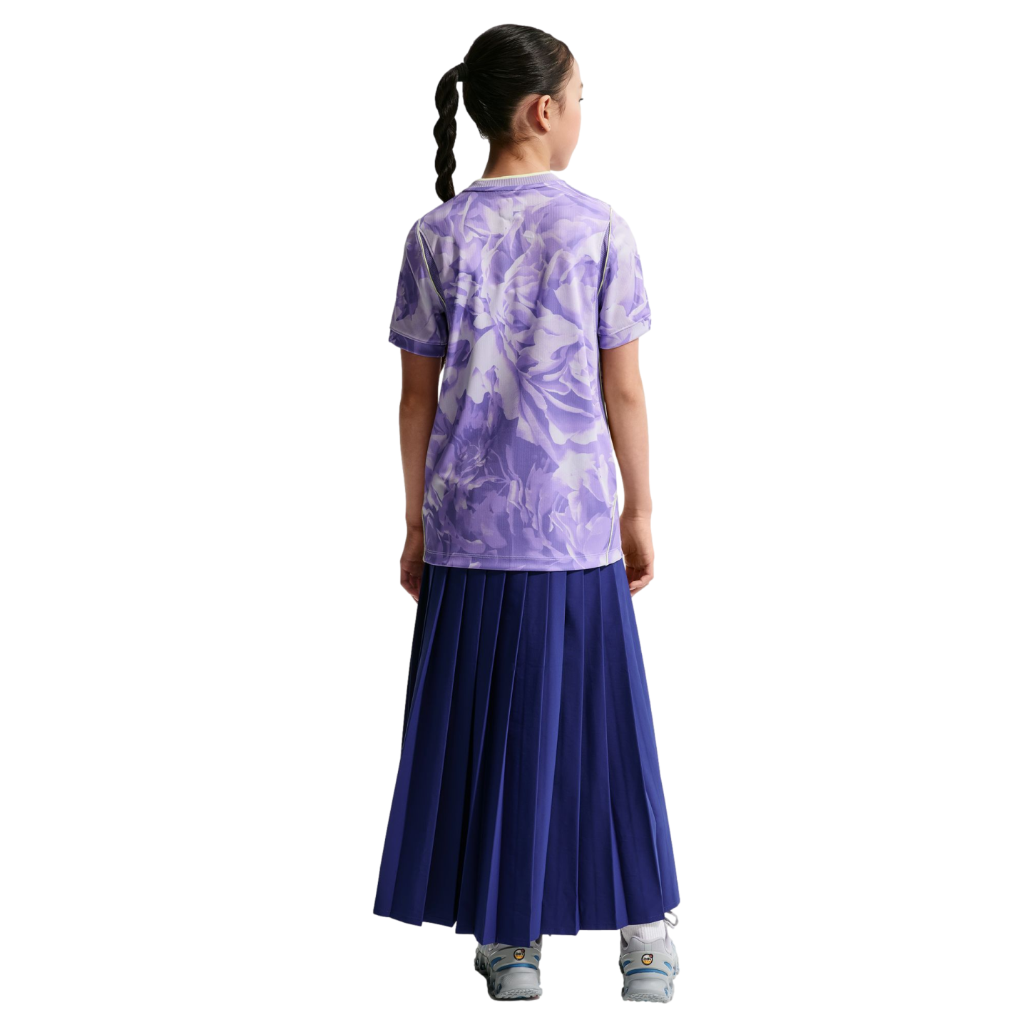Back view of a girl with a braided ponytail wearing a purple floral t-shirt and a long navy pleated skirt against a black background.
