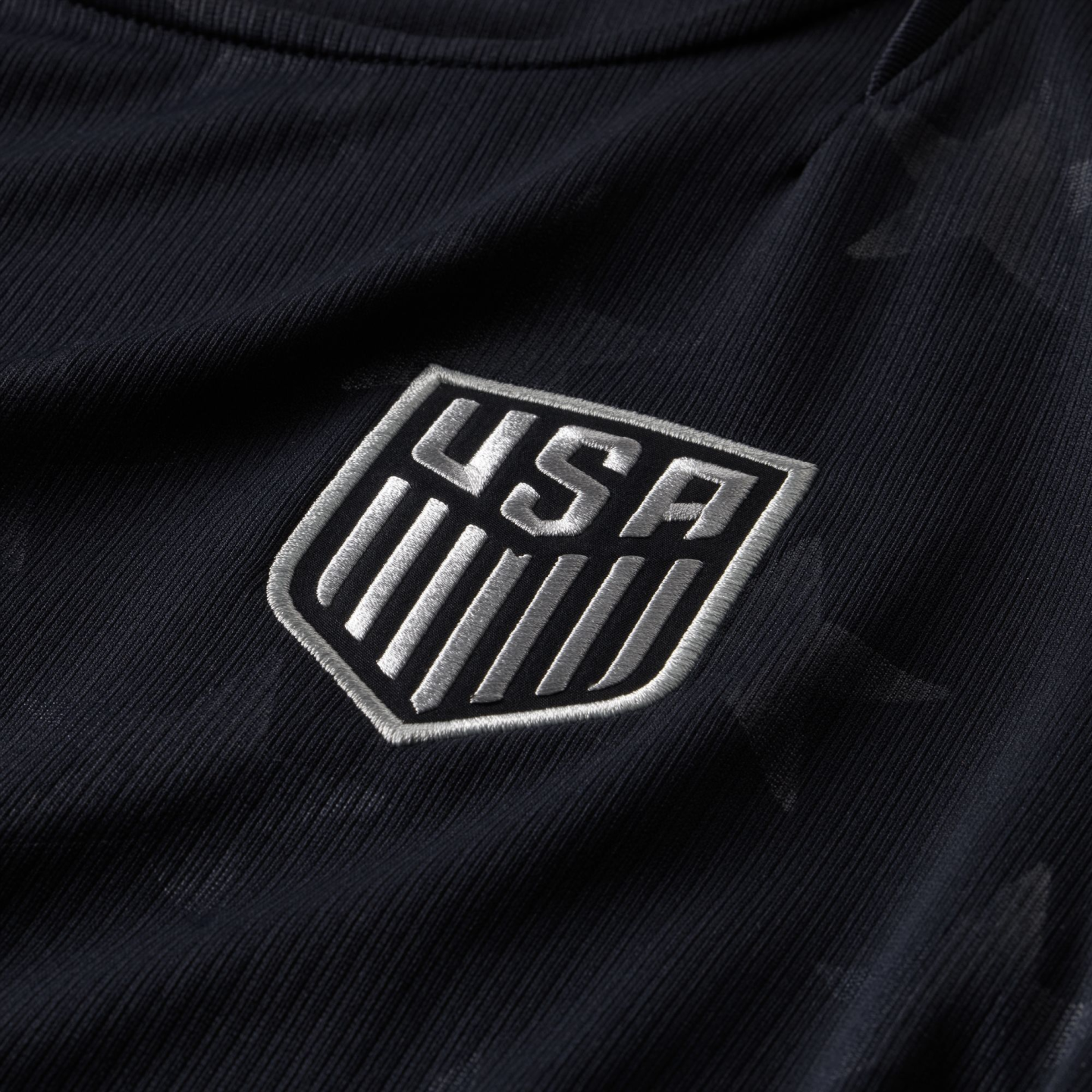 A close-up of a dark ribbed jersey featuring an embroidered silver U.S. Soccer Federation shield crest with "USA" and vertical stripes.