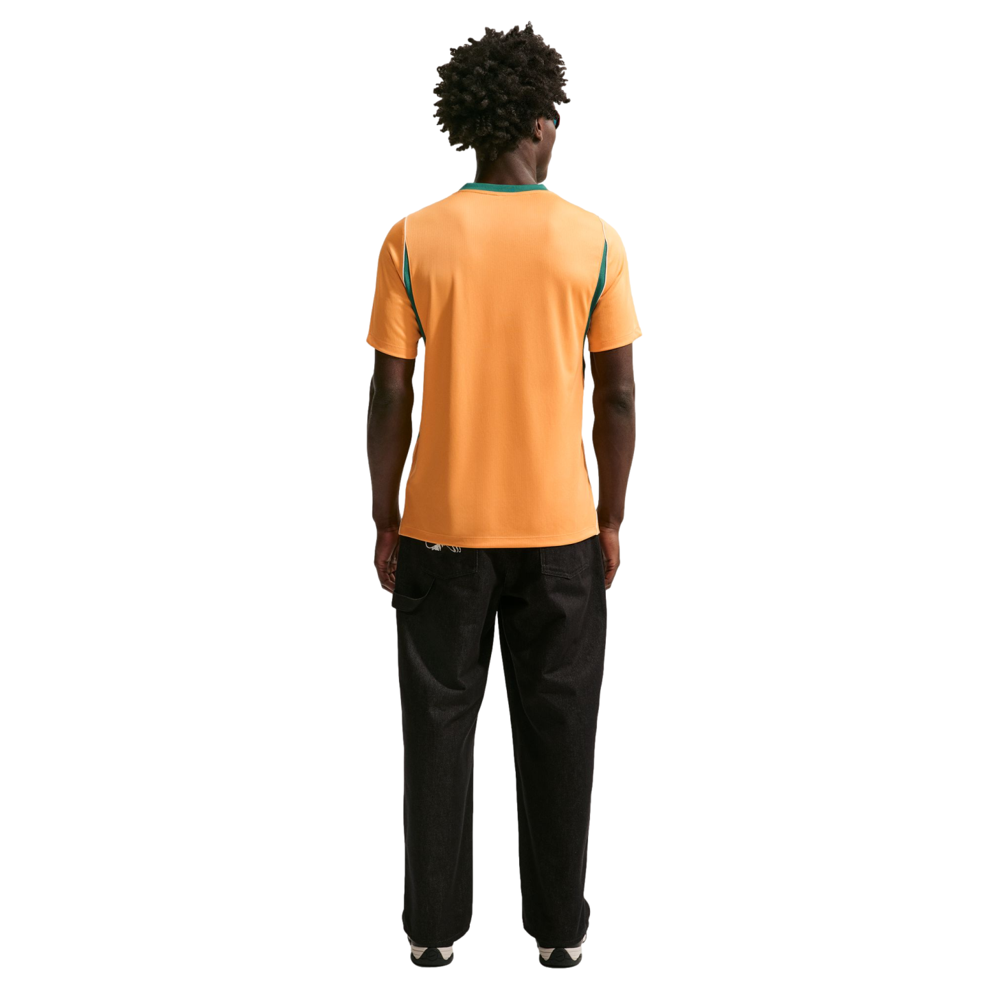 Back view of a person with dark curly hair wearing an orange shirt with green trim and black pants against a black background.