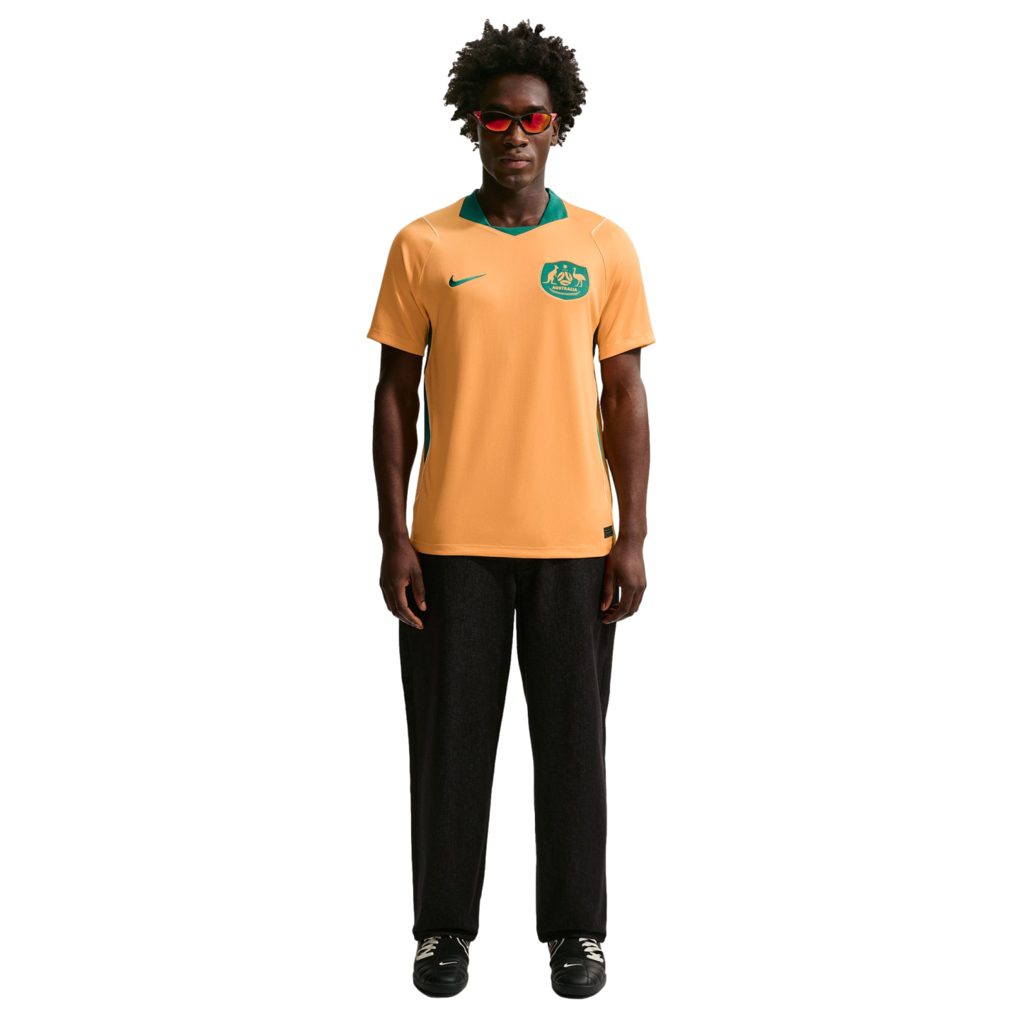A man with curly hair and red sunglasses wears an orange and green Australia soccer jersey with black pants, standing against a solid black background.