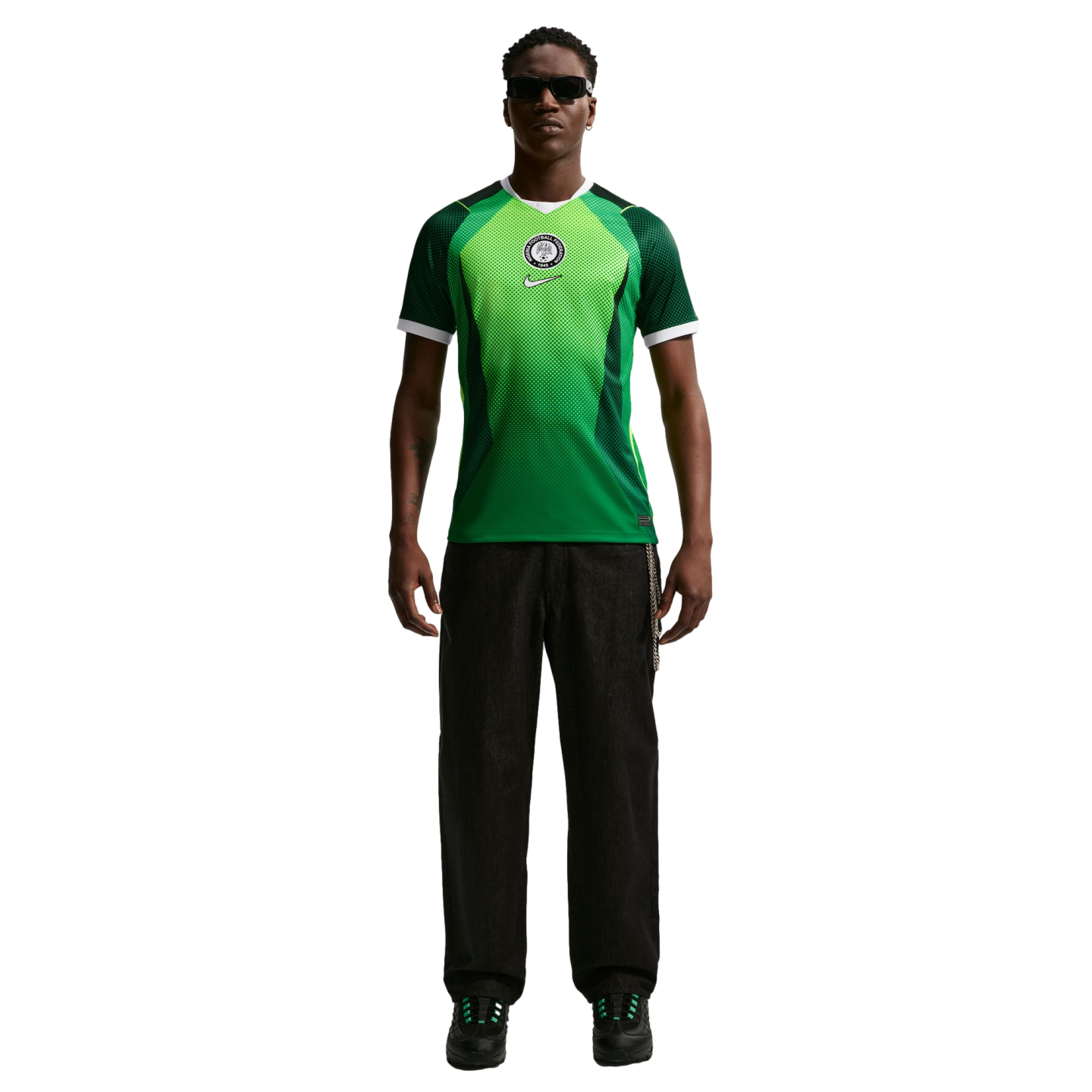 A man wearing a green and white Nigeria football jersey, black pants, and sunglasses, standing against a black background.
