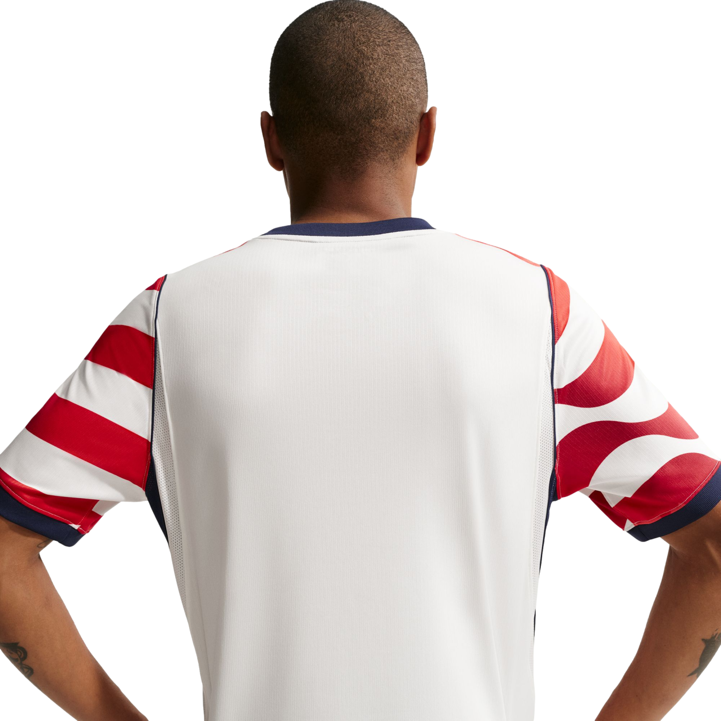 Rear view of a man with a buzz cut wearing a white jersey with red-and-white patterned sleeves and navy trim, standing with hands on hips against a black background.