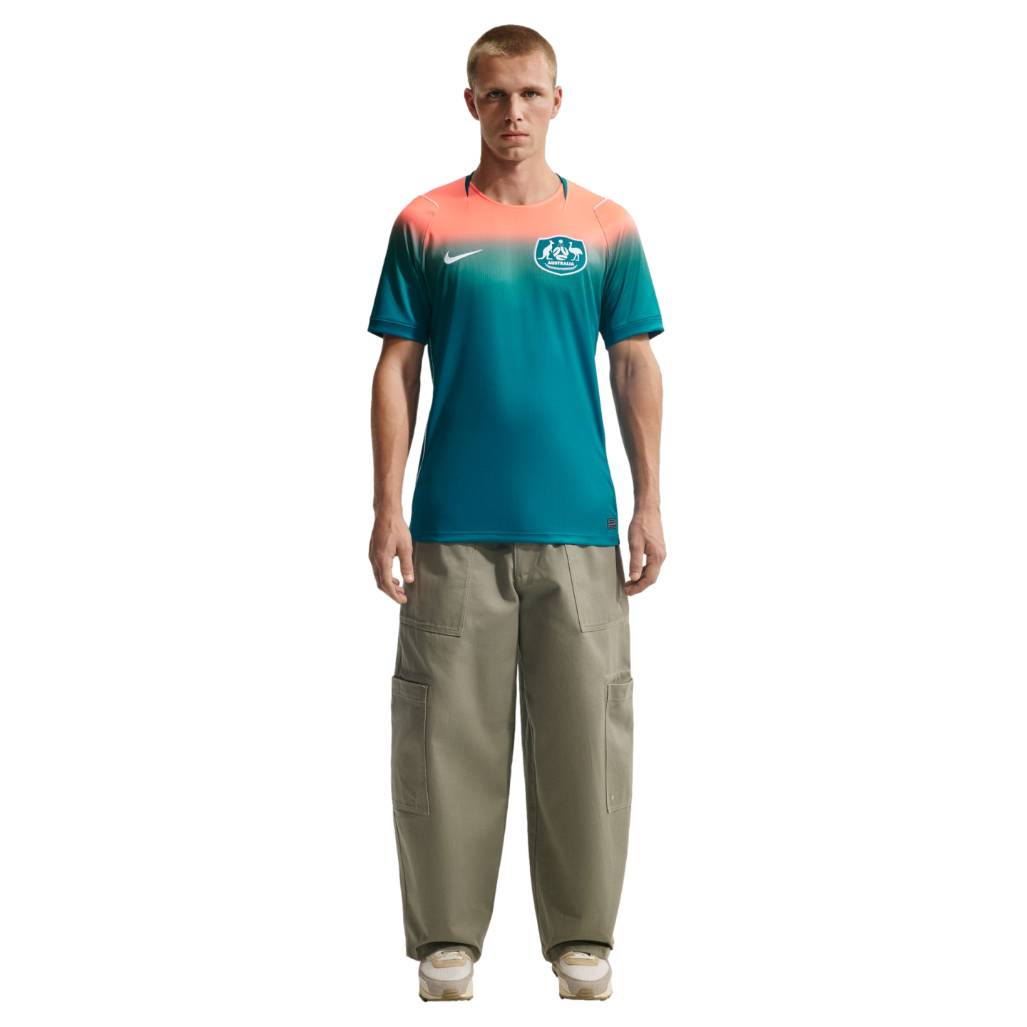 A man standing against a black background wearing a Nike Australia national team jersey with a peach-to-teal gradient and oversized olive green cargo pants.