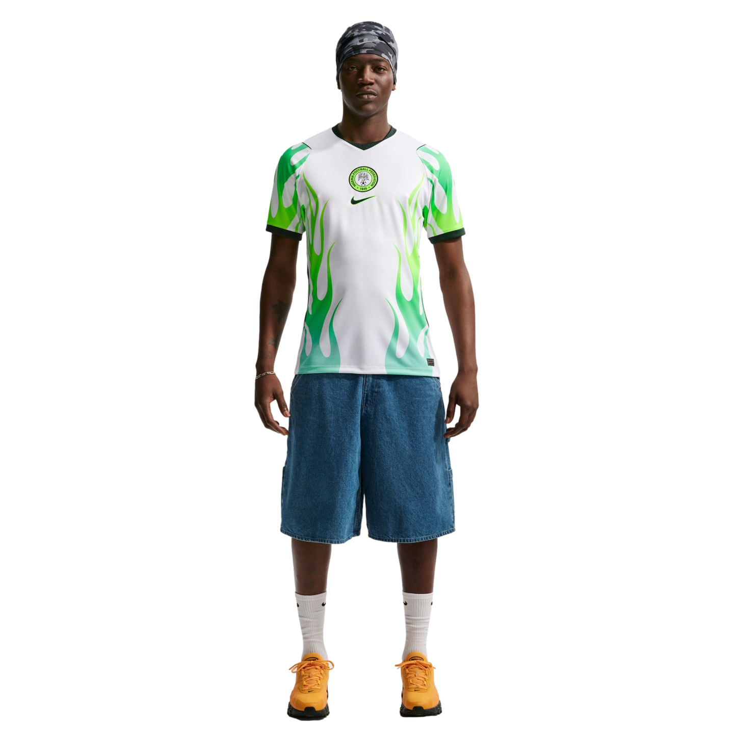 A man wearing a white and green flame-patterned soccer jersey, denim shorts, and orange sneakers stands against a black background.