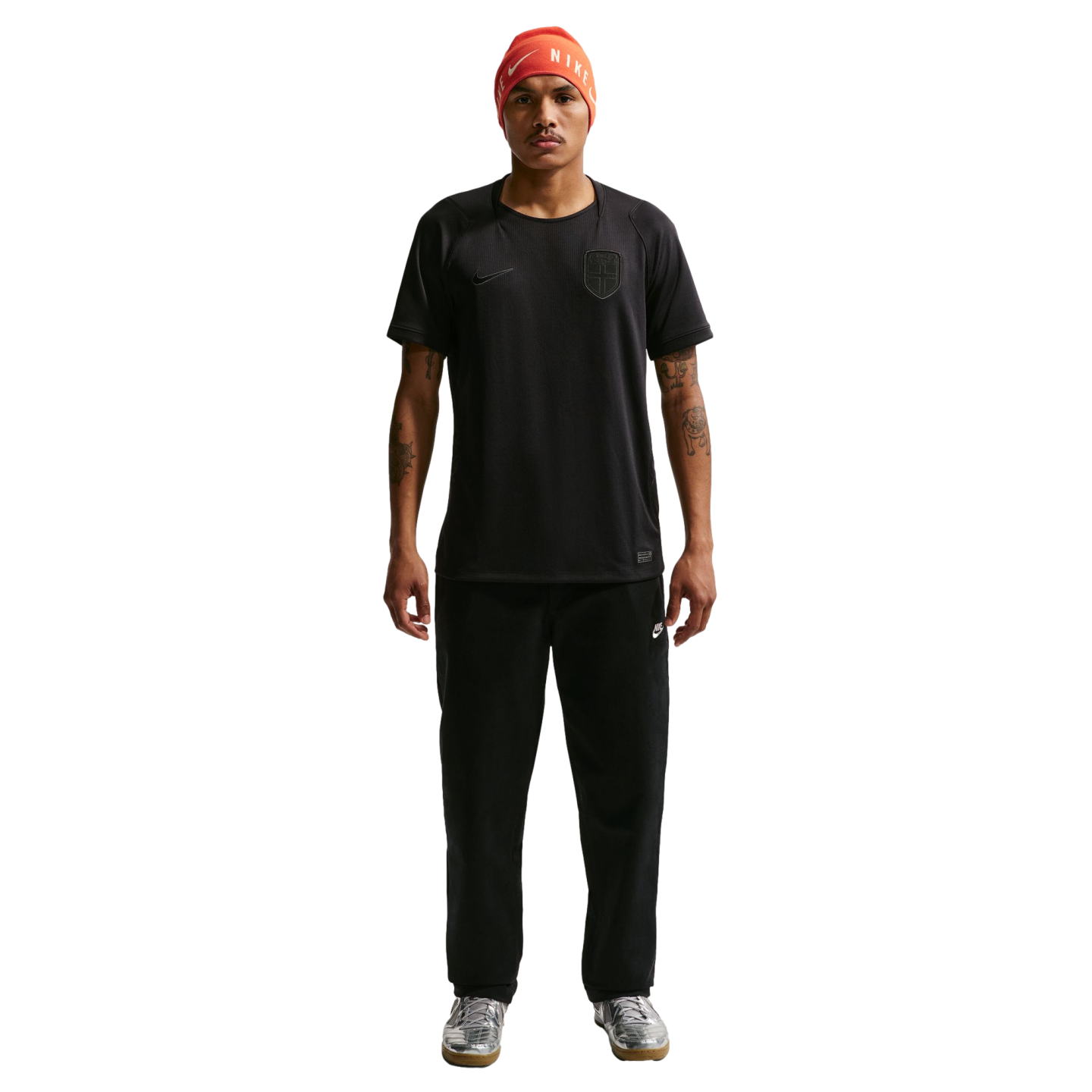 A man wearing an orange Nike beanie, a black t-shirt and sweatpants, and silver sneakers stands against a black background.