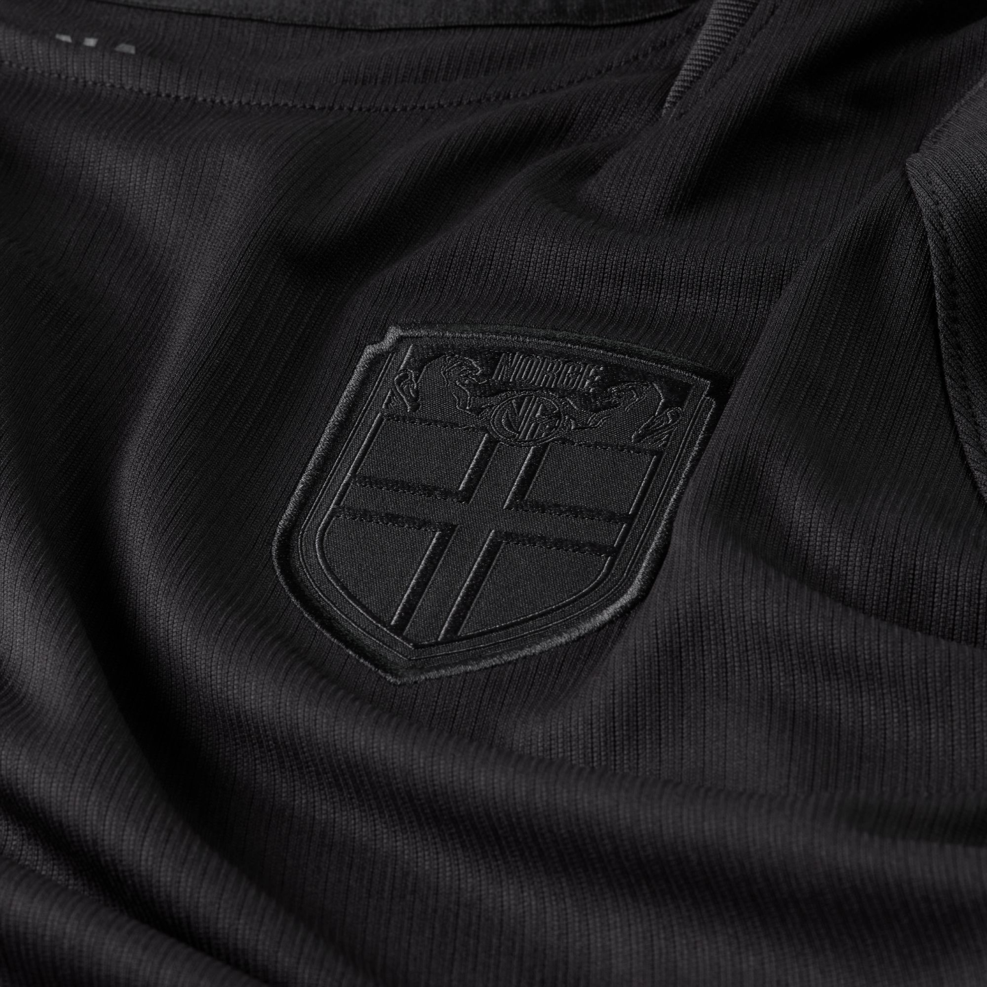 Close-up of a black textured jersey featuring an all-black embossed crest with a cross and the word "NORGE."