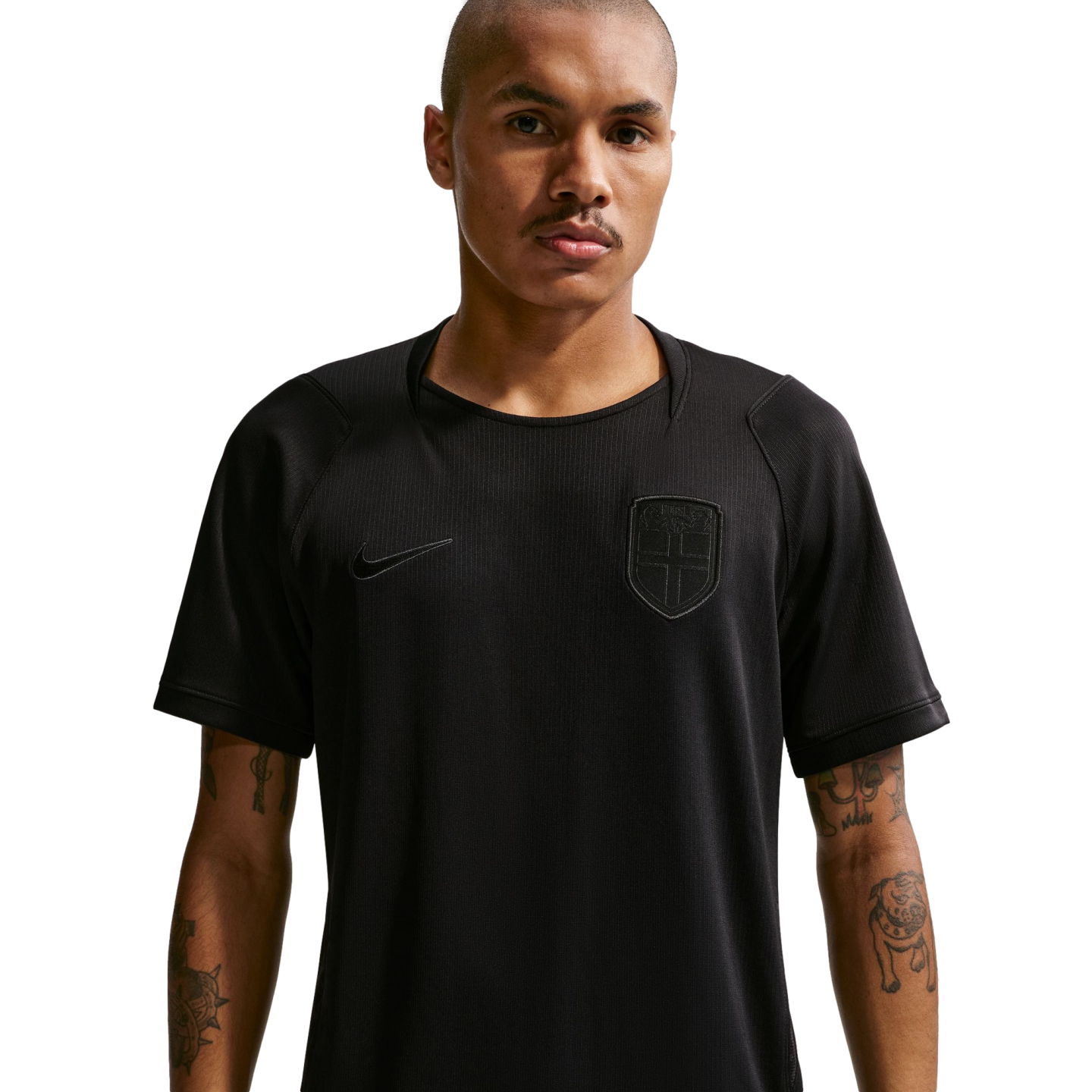 A man with a buzz cut and forearm tattoos wears a black Nike athletic shirt with a subtle monochromatic shield crest, standing against a dark background.