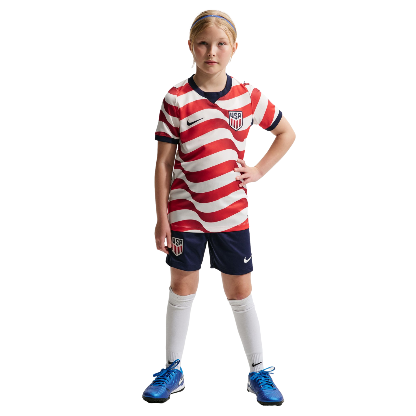 A young girl in a red and white wavy striped USA soccer uniform, navy shorts, and bright blue cleats, standing against a solid black background.