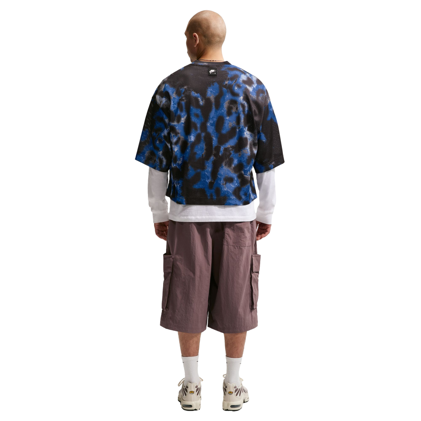 Rear view of a man wearing a blue and black patterned mesh tee layered over a white long-sleeve shirt, mauve cargo shorts, and white sneakers.