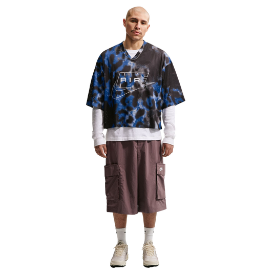 A man wears an oversized blue and black patterned Nike jersey, white long-sleeve shirt, baggy brown cargo shorts, and cream sneakers against a white background.
