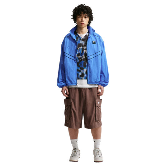 A man with curly hair poses in a blue windbreaker, a patterned top, brown cargo shorts, and sneakers against a plain white background.