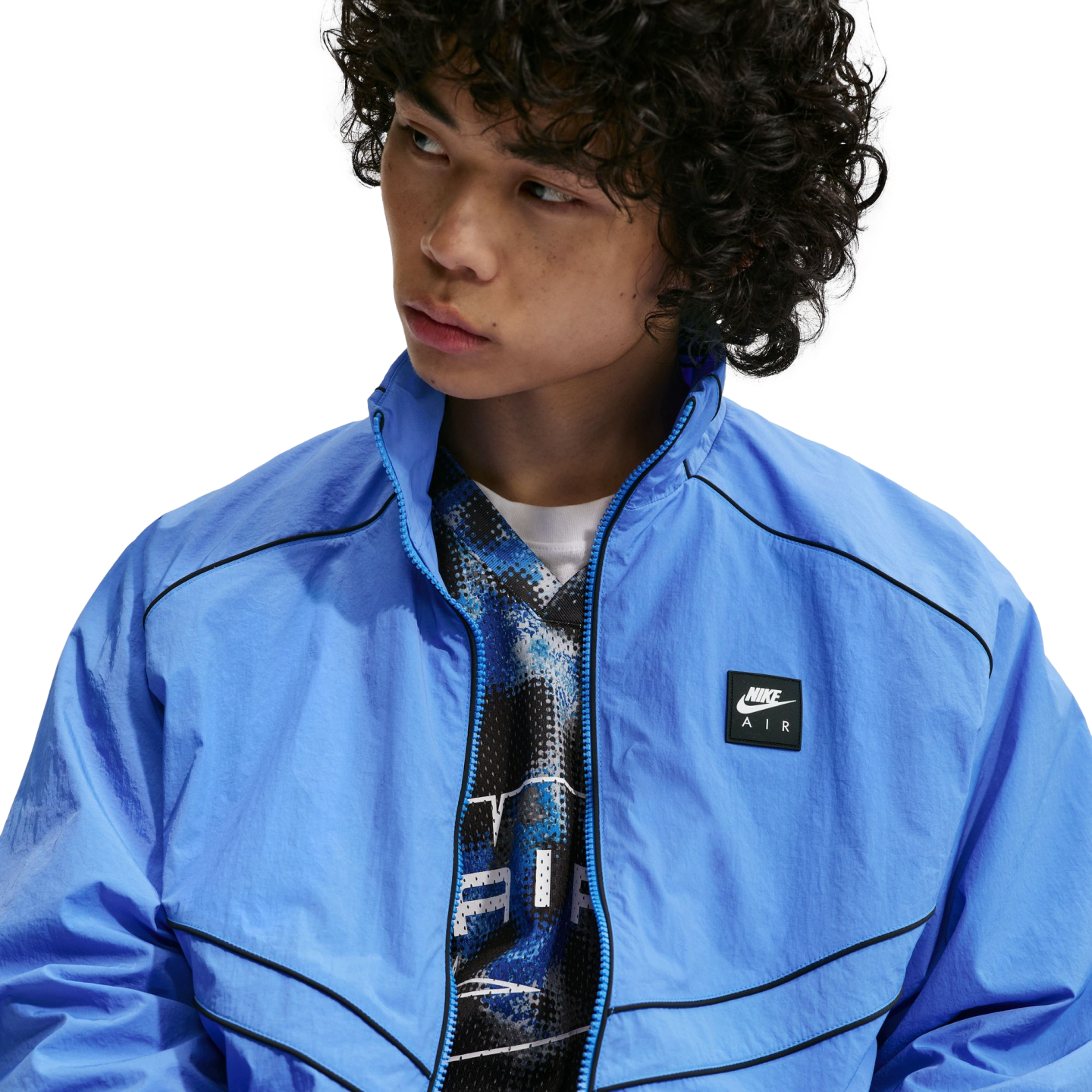 Young man with dark curly hair wearing a bright blue Nike Air windbreaker jacket over a patterned shirt, looking to the side against a white background.