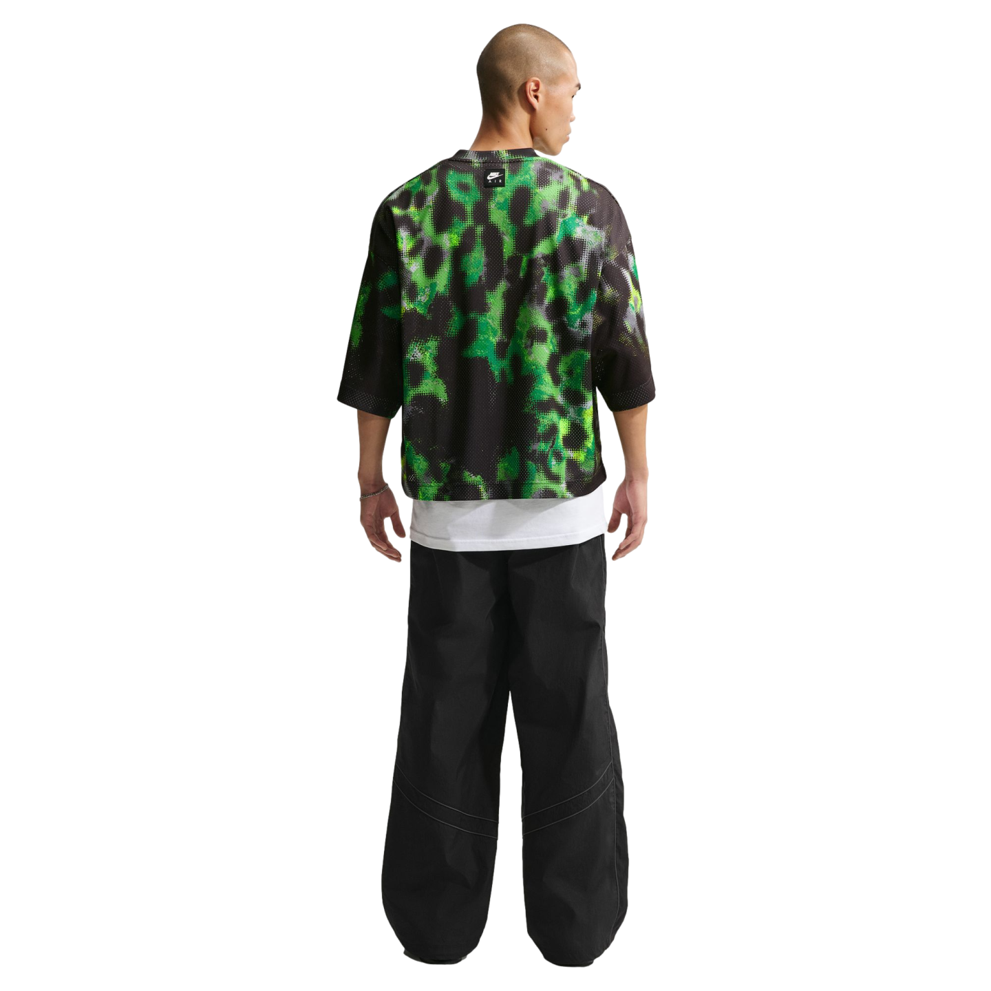 Back view of a person wearing a green and black mesh top over a white tee and baggy black trousers against a white backdrop.