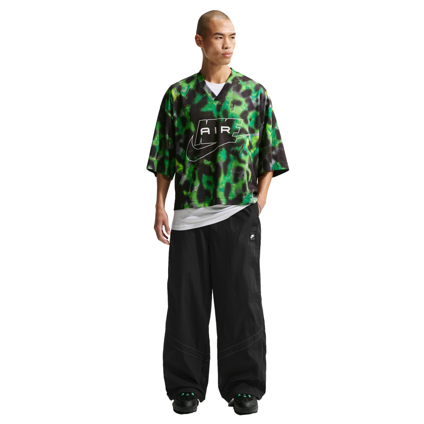 A man with a shaved head wearing a patterned green and black Nike mesh jersey, baggy black pants, and black sneakers against a white background.