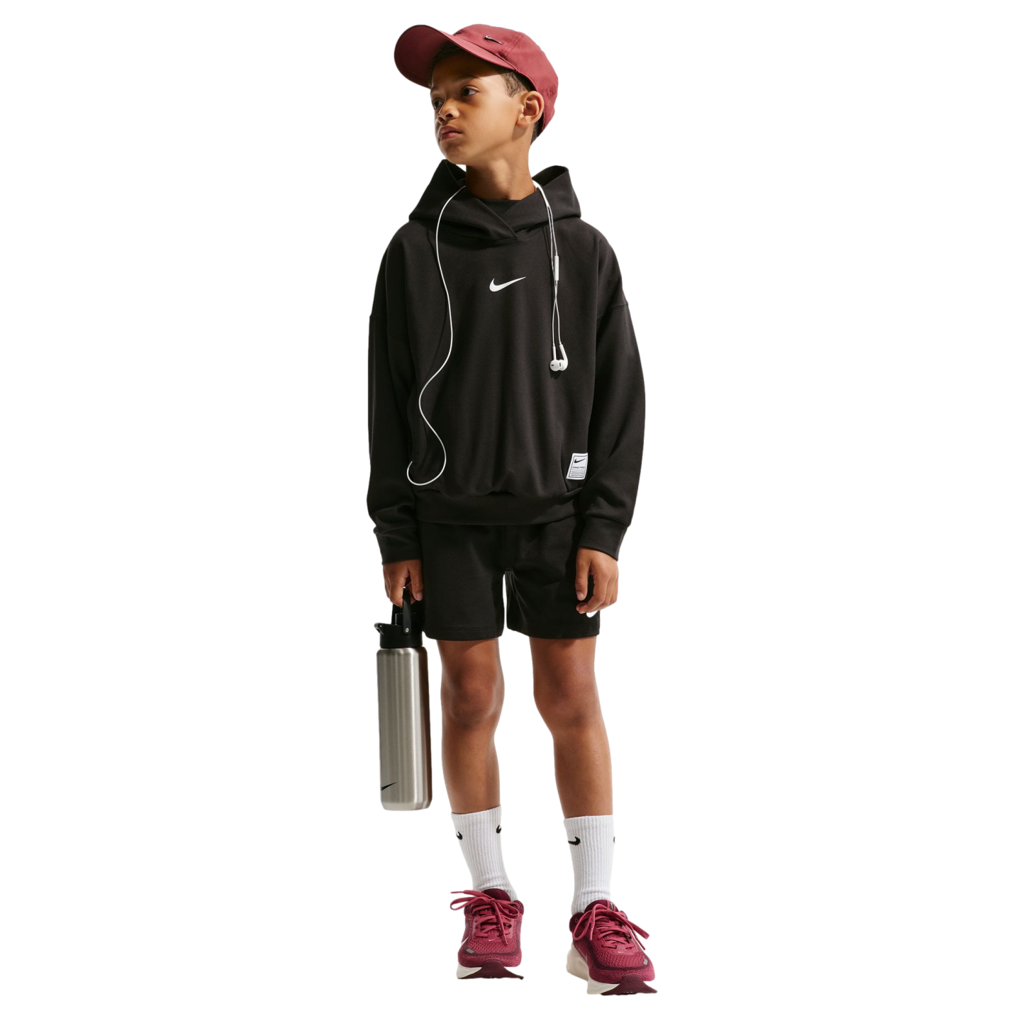 A young boy in a black Nike hoodie, black shorts, maroon cap, and maroon sneakers stands against a black background, holding a silver water bottle and wearing wired earphones.