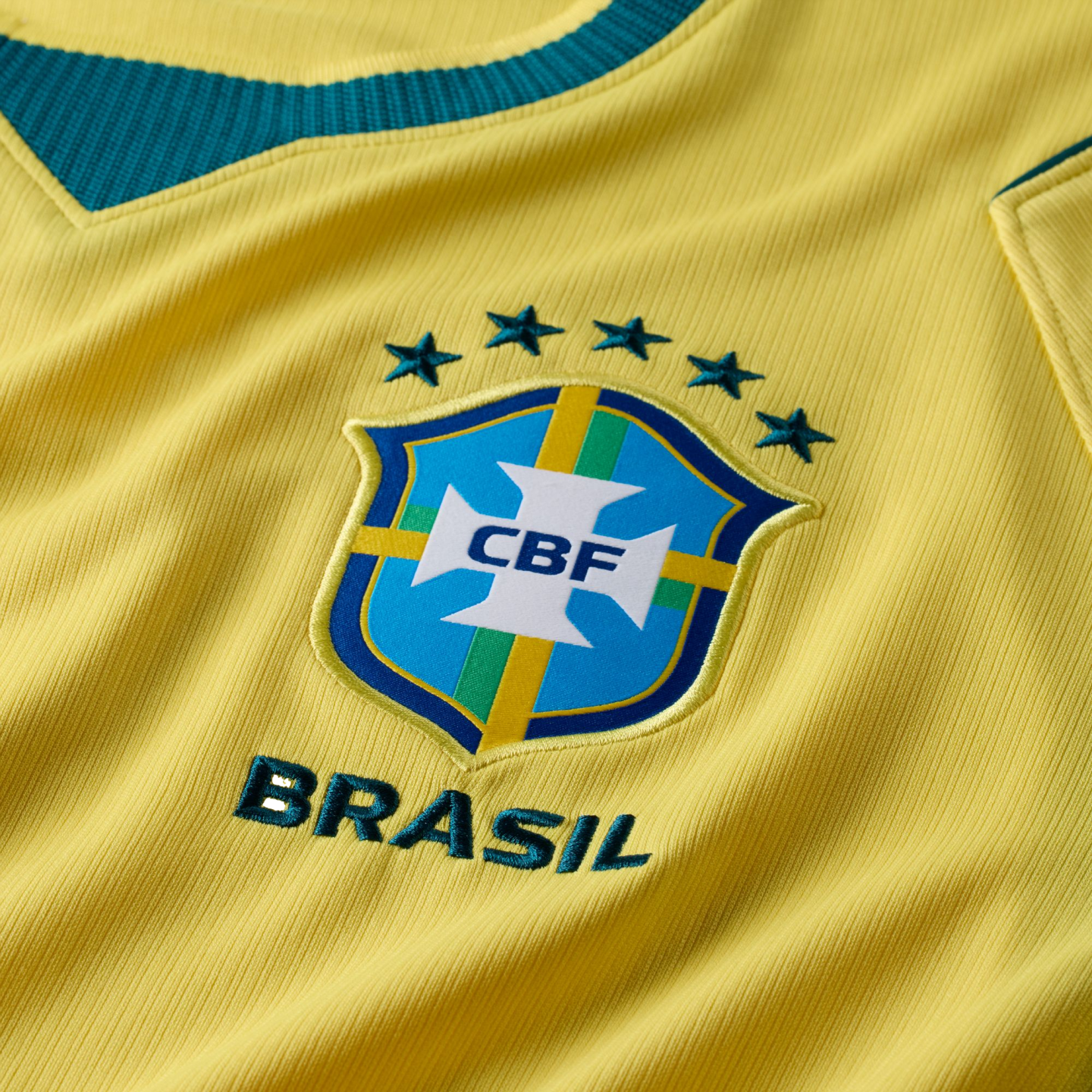 A close-up shot of a yellow Brazilian national soccer team jersey, featuring the embroidered CBF crest in the center. Above the crest are five green stars arranged in an arc, and the word "BRASIL" is embroidered in green underneath. A portion of the dark 