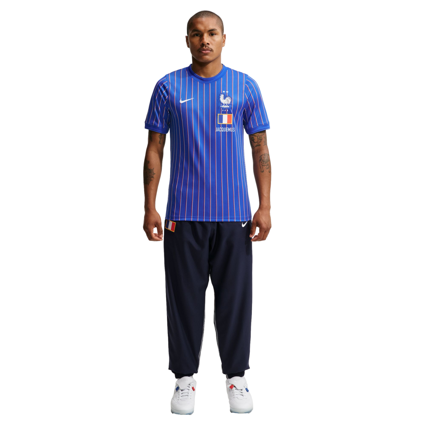 A man wearing a blue pinstriped Nike x Jacquemus France soccer jersey, navy track pants, and white sneakers stands against a plain white background.