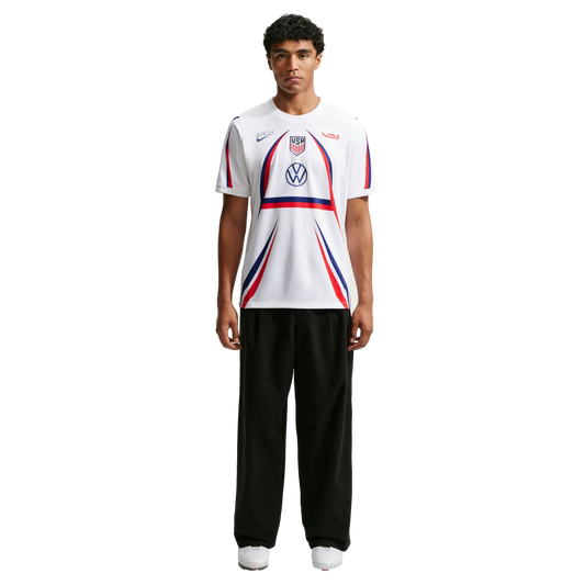 A man stands wearing a white USA soccer jersey with red and blue accents, black wide-leg trousers, and white sneakers against a plain white background.
