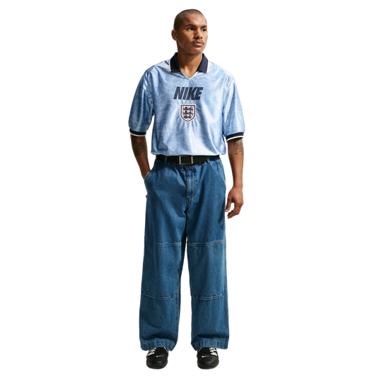 A man models a light blue Nike England jersey tucked into baggy wide-leg blue jeans and black sneakers against a plain white background.