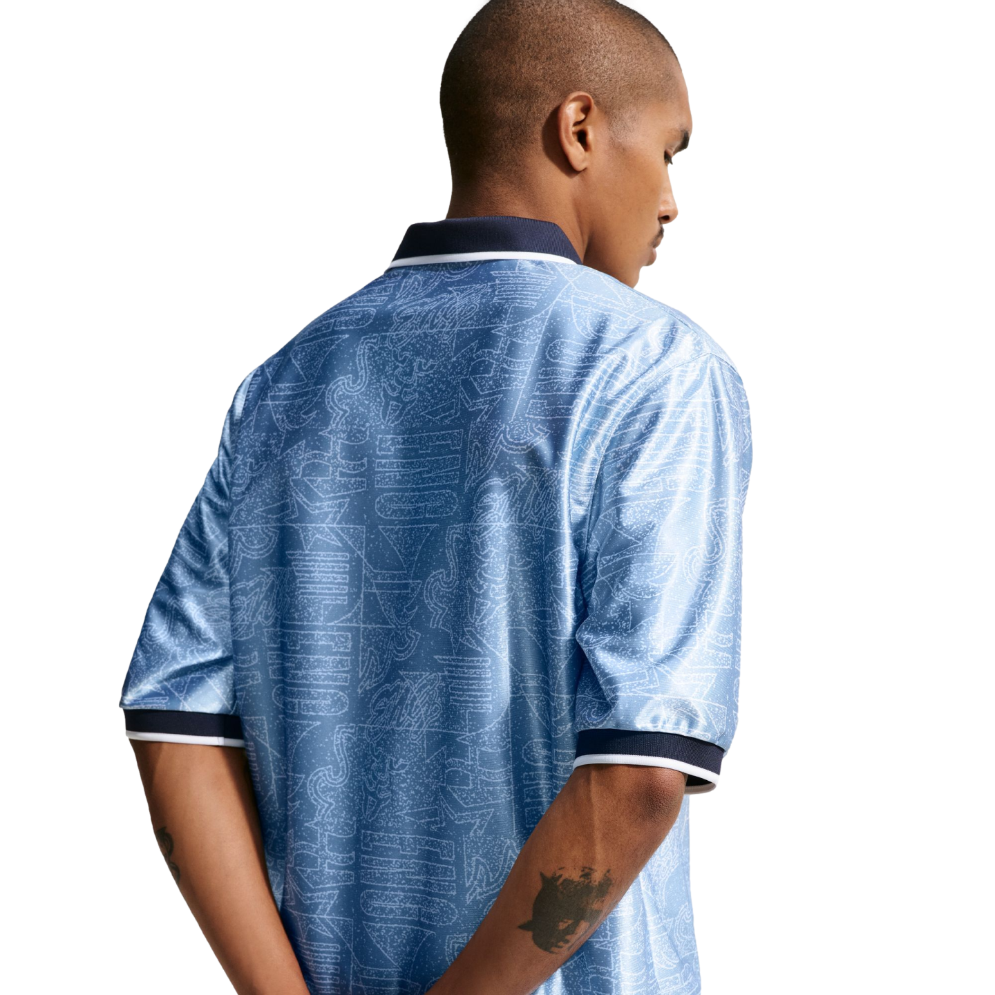 Back view of a man in a patterned light blue polo shirt with navy and white trim, standing against a white background with his hands behind his back.