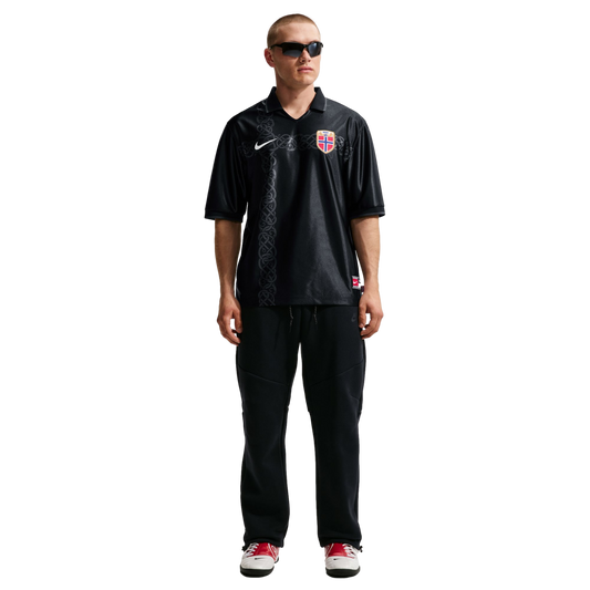 A man modeling a black Nike Norway football jersey with a Nordic knot pattern, black sweatpants, sunglasses, and red and white sneakers against a white background.