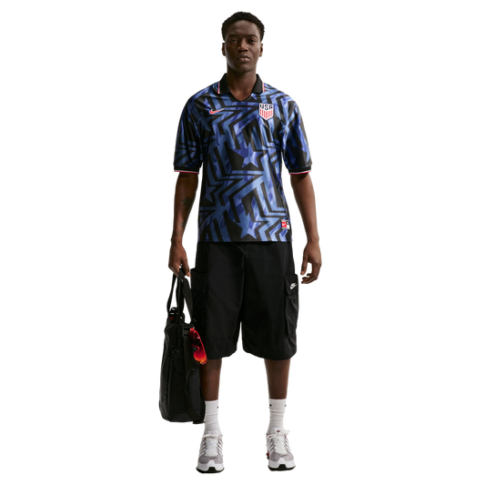 A Black man wearing a blue patterned USA soccer jersey, black cargo shorts, and sneakers stands holding a black bag against a white background.