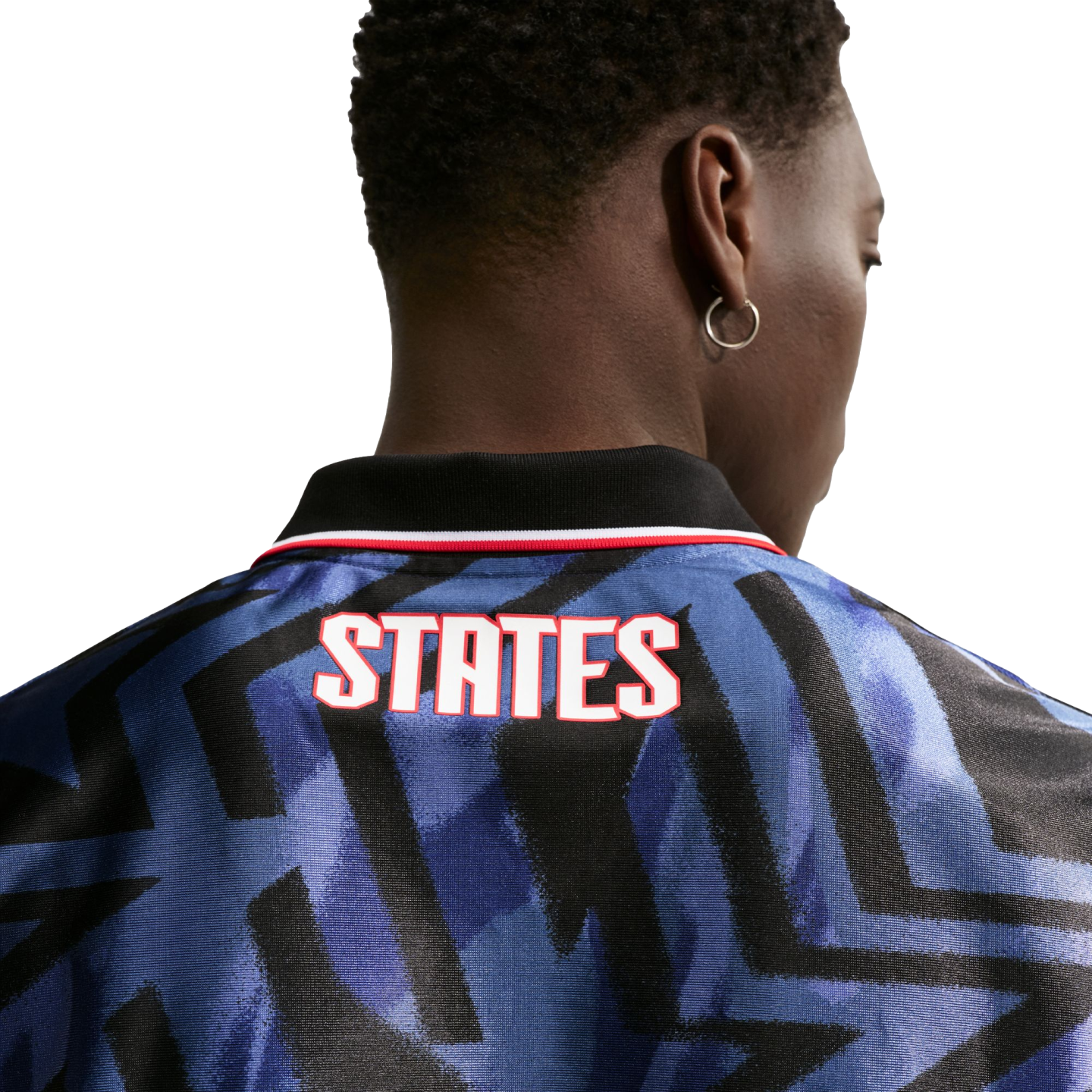 Rear view of a person with short dark hair wearing a blue patterned jersey with "STATES" in white and red text on the back. They are wearing a silver hoop earring.