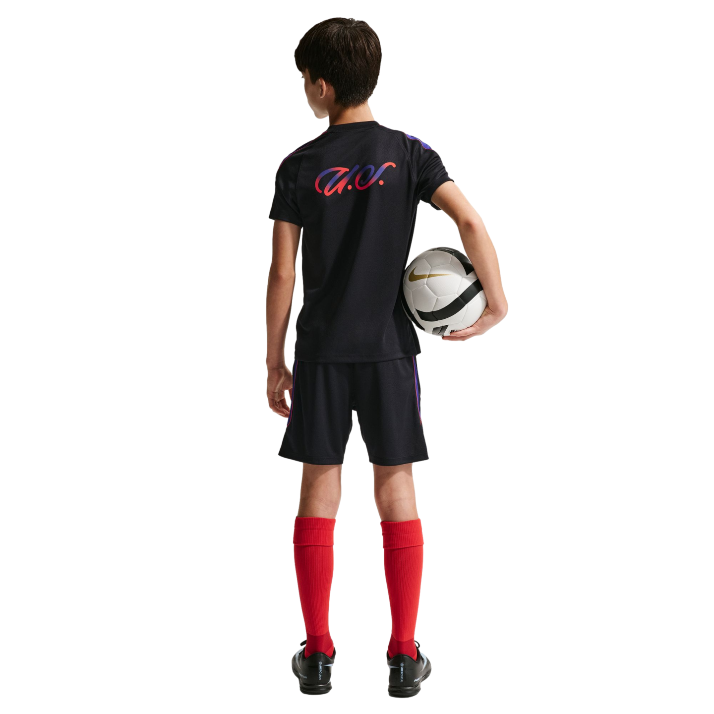 Back view of a young boy in a black soccer kit and red socks, holding a white and black soccer ball against a solid black background.