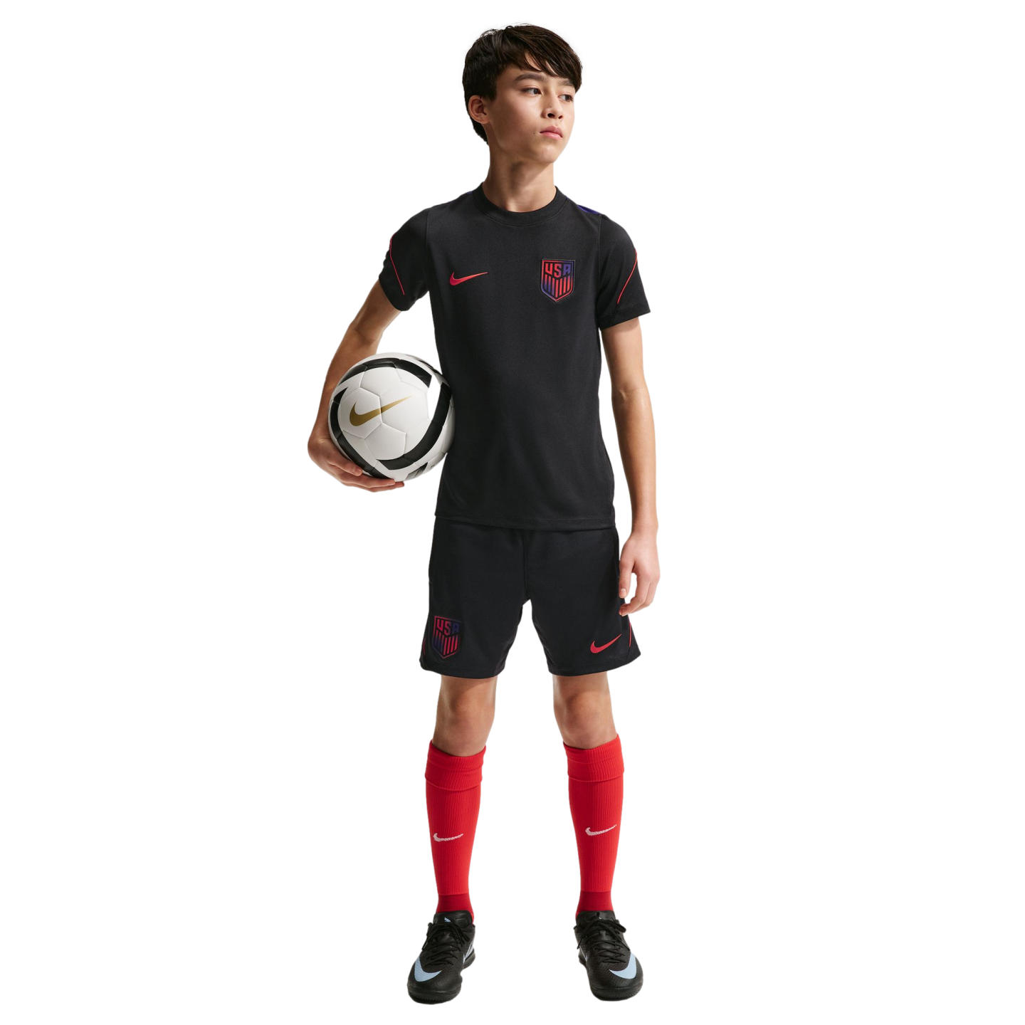 A young boy in a black USA soccer kit and red socks holds a soccer ball against a solid black background.