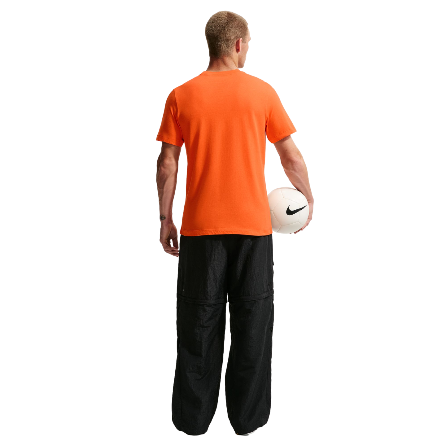A rear view of a man standing against a black background, wearing a bright orange T-shirt and baggy black pants. He holds a white Nike soccer ball under his left arm.