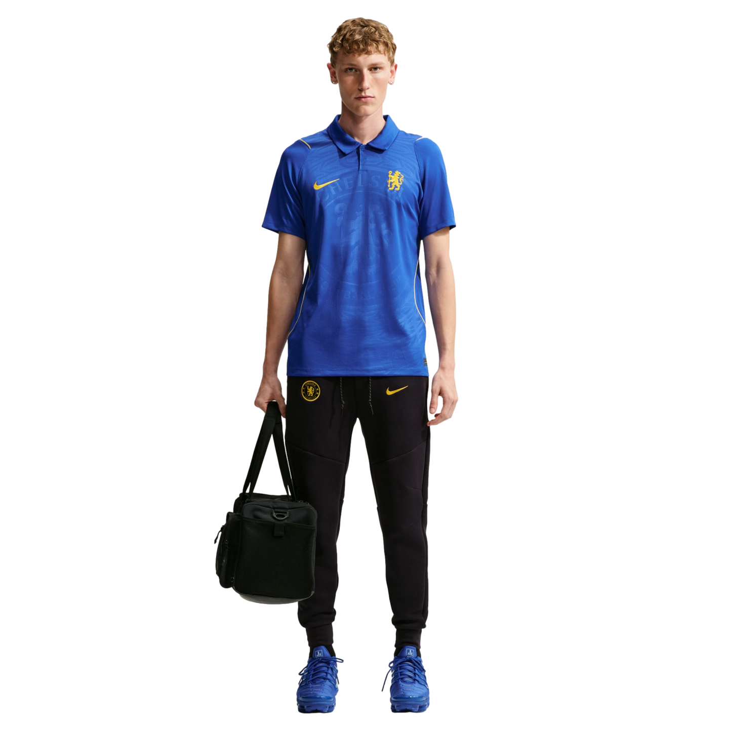 A person wearing a blue Chelsea FC polo shirt, black Nike joggers, and blue sneakers, holding a black gym bag.