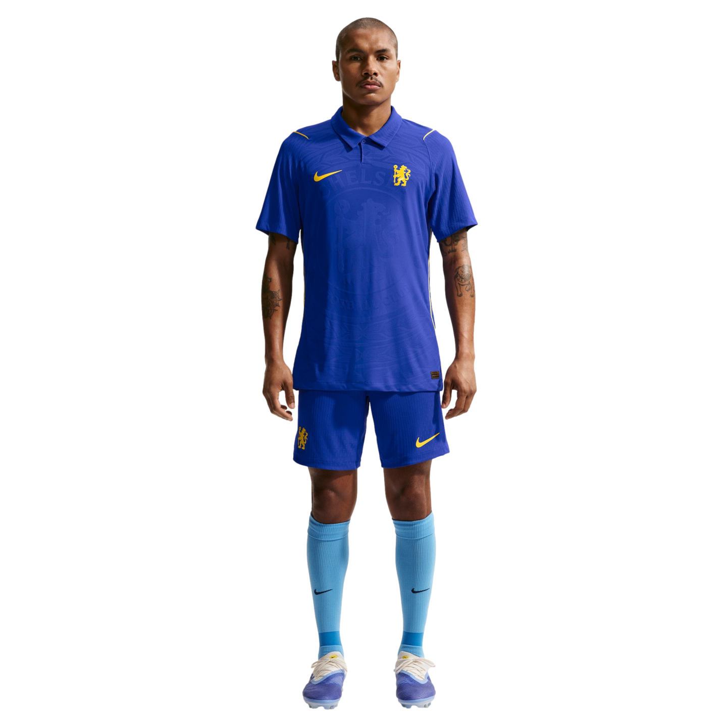 A man stands against a white background wearing a blue Nike Chelsea FC football kit with a polo-style jersey, matching blue shorts, and light blue socks.